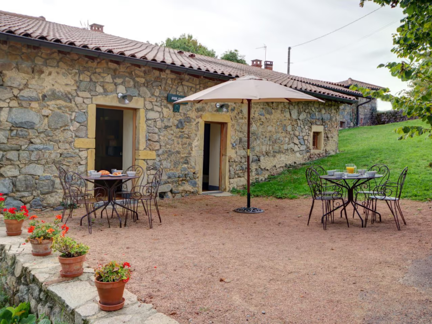 Une terrasse en pierre avec des tables et des chaises en fer forgé, un parasol blanc, devant une maison en pierre avec une porte et deux fenêtres, entourée d'arbres et de végétation.