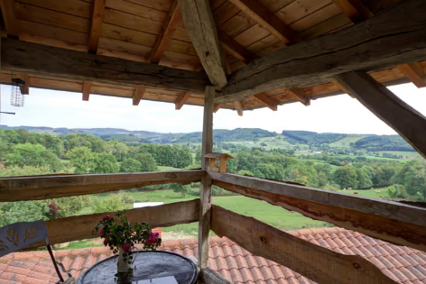 Vue panoramique depuis une terrasse en bois avec une table, des chaises et des fleurs, surplombant une campagne verdoyante avec des collines et des champs.