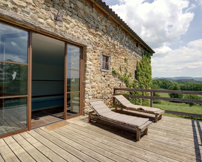 Terrasse en bois avec deux transats en face d'une maison en pierre avec fenêtre et vue sur la campagne