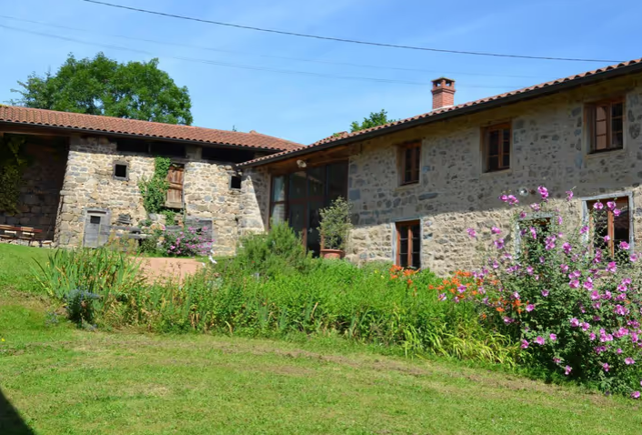 Maison en pierre avec jardin fleuri et arbres sous un ciel bleu.