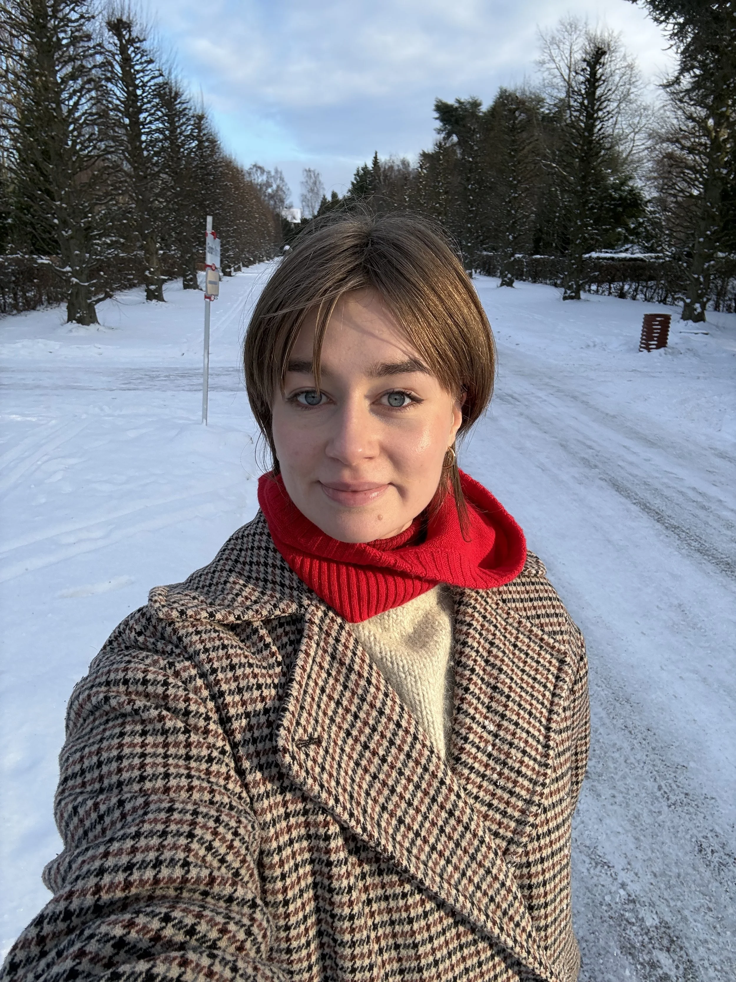 A woman taking a selfie outdoors in a snowy park with snow-covered trees and a cloudy sky in the background.