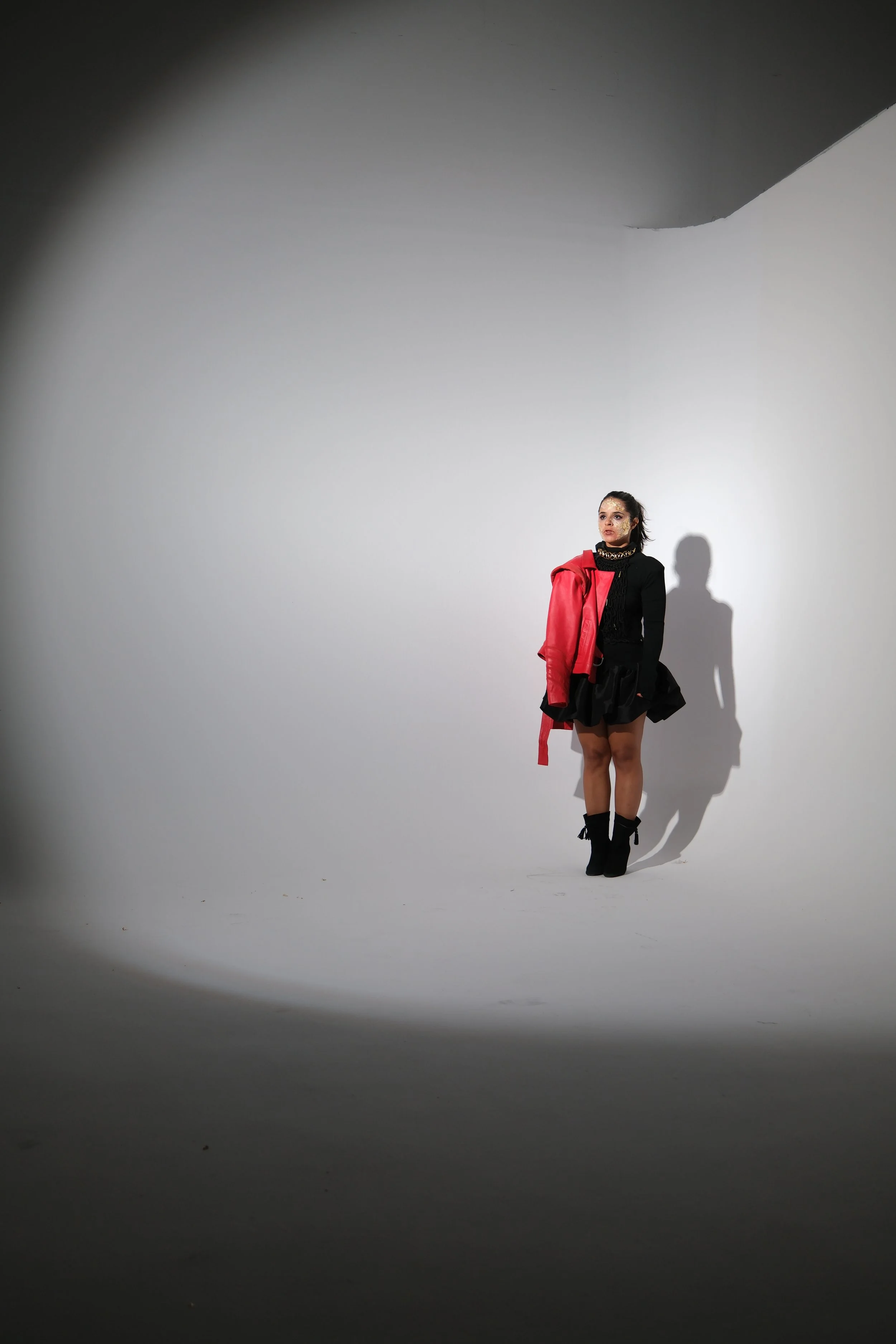 Une femme debout dans un studio blanc, portant une veste rouge à une épaule, une jupe noire à volants, des bottes noires hautes, avec un maquillage audacieux et une expression sérieuse.