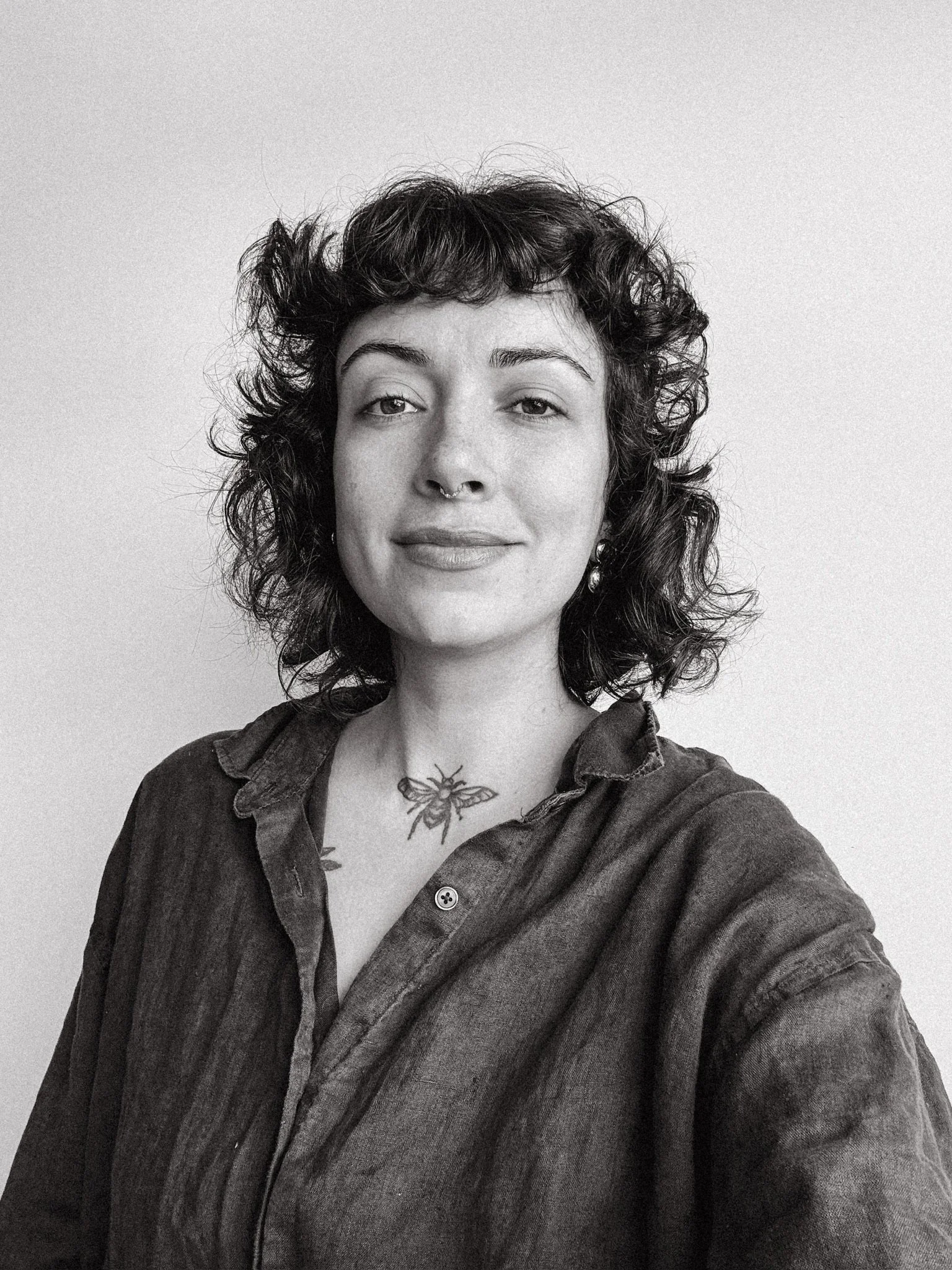 Retrato preto e branco de uma mulher com cabelo curto, cacheado e escuro, usando uma camisa de tecido escuro com colarinho aberto, exibindo uma tatuagem de uma abelha no colo, sorrindo levemente e olhando para a câmera.