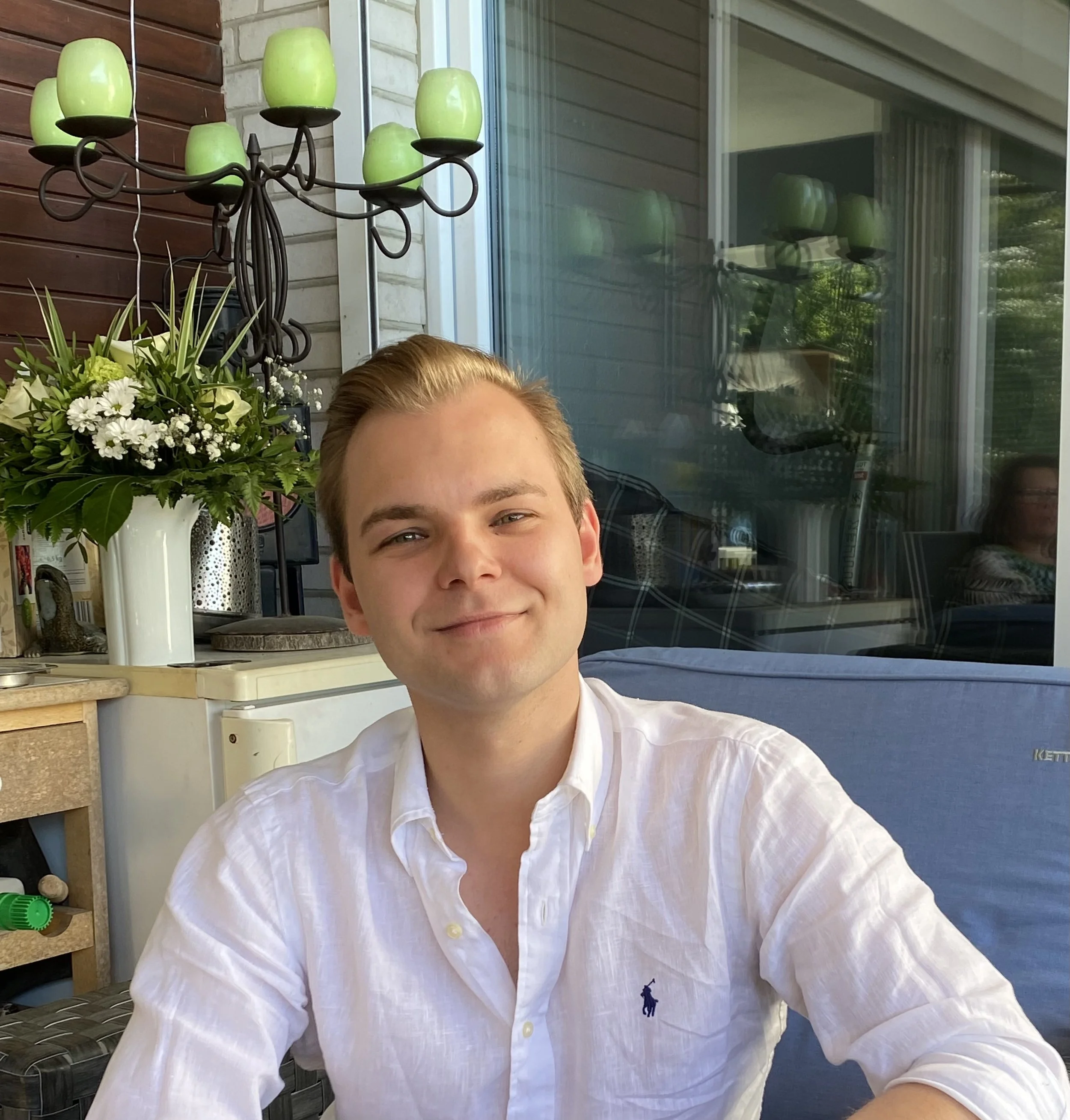 Nahaufnahme eines lächelnden jungen Mannes mit blonden Haaren in einem weißen Hemd, sitzend auf einer Terrasse mit Blumen und einem Garten im Hintergrund.