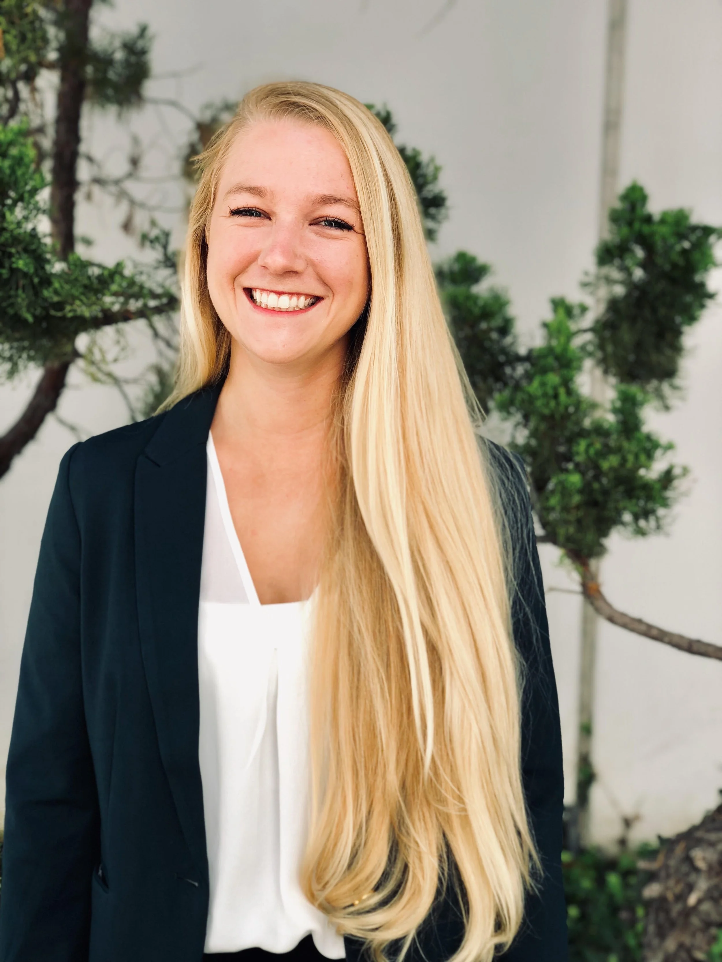 A smiling woman with long blonde hair, wearing a black blazer and white blouse, standing outdoors with trees in the background.