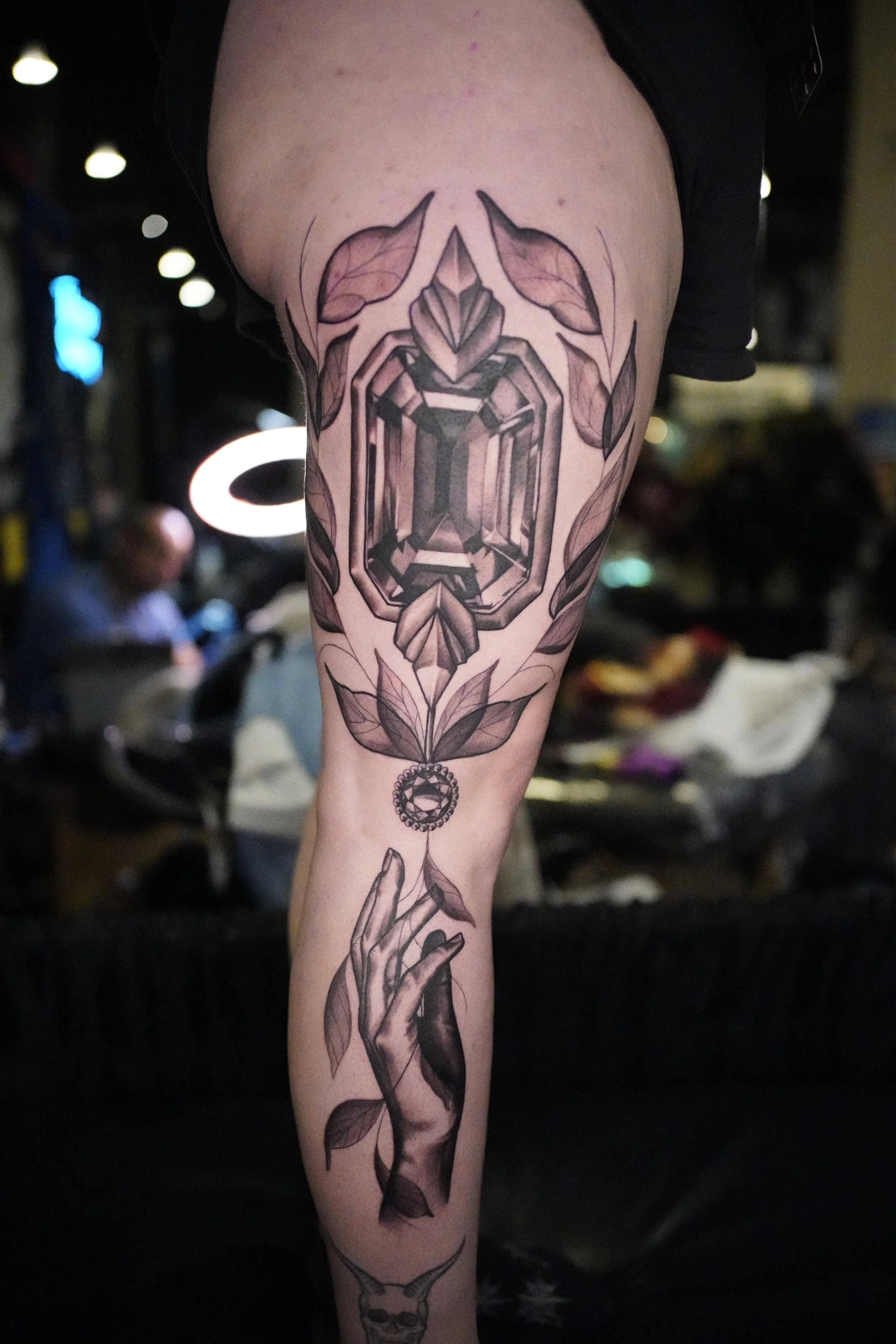 Tattoo of a large geometric gem surrounded by leaves and a hanging ornament on a person's arm.