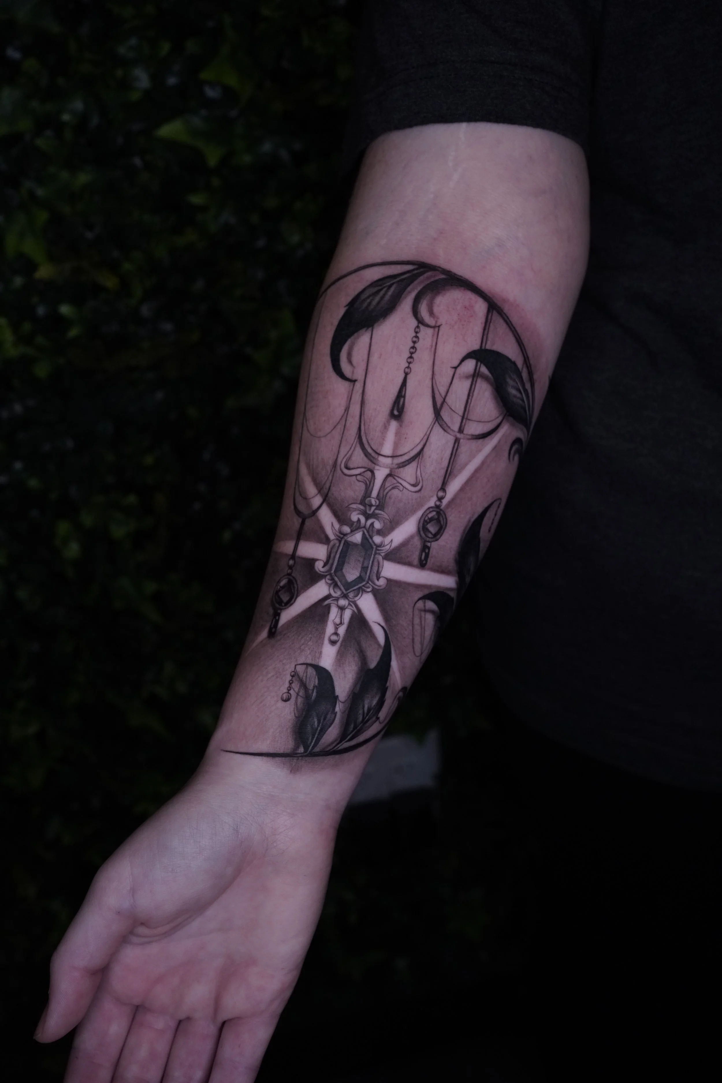 A person's forearm with a black and gray tattoo of feathers, chains, and jewelry designs.