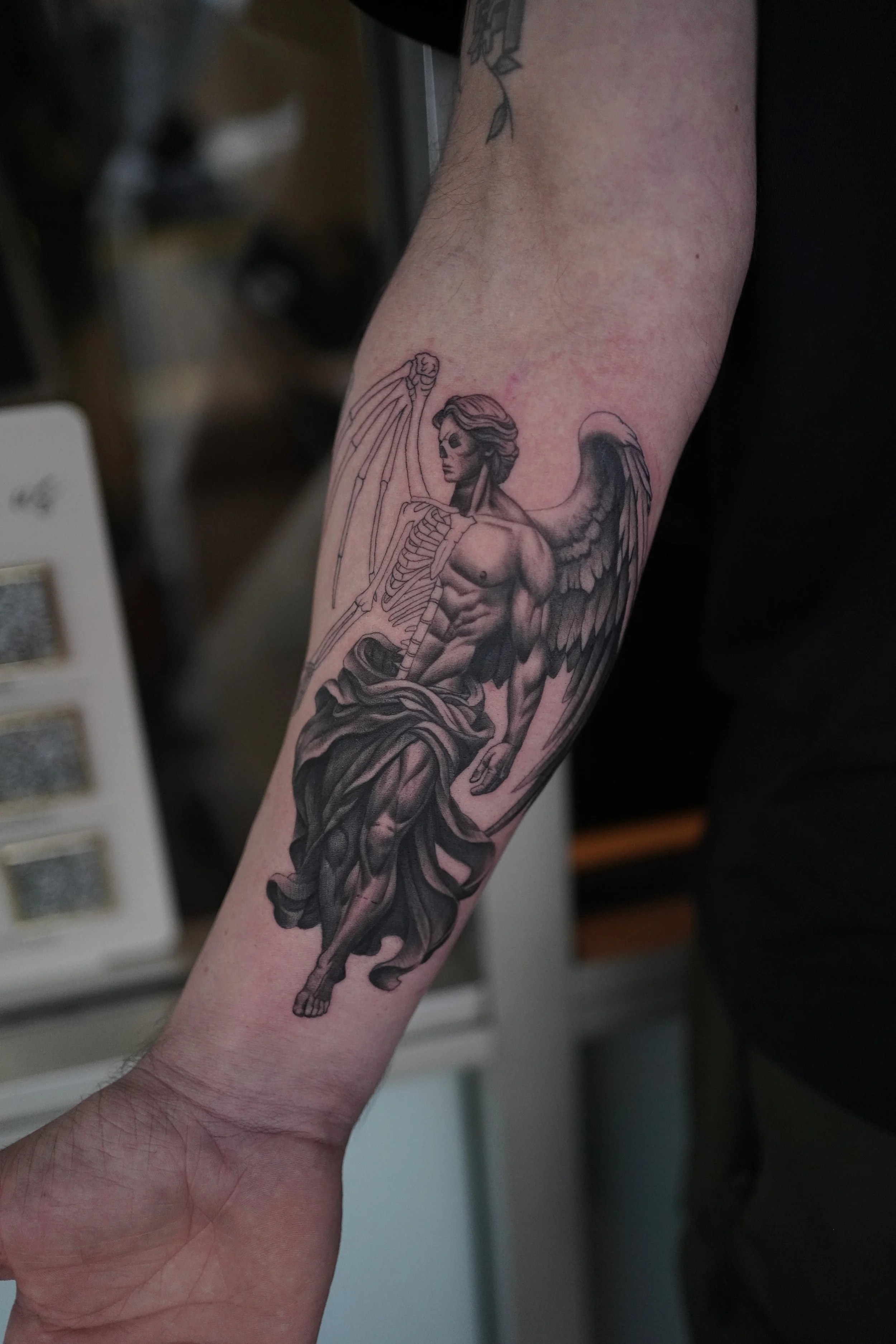 Tattoo of a skeletal angel with wings and a muscular male body on a person's forearm.