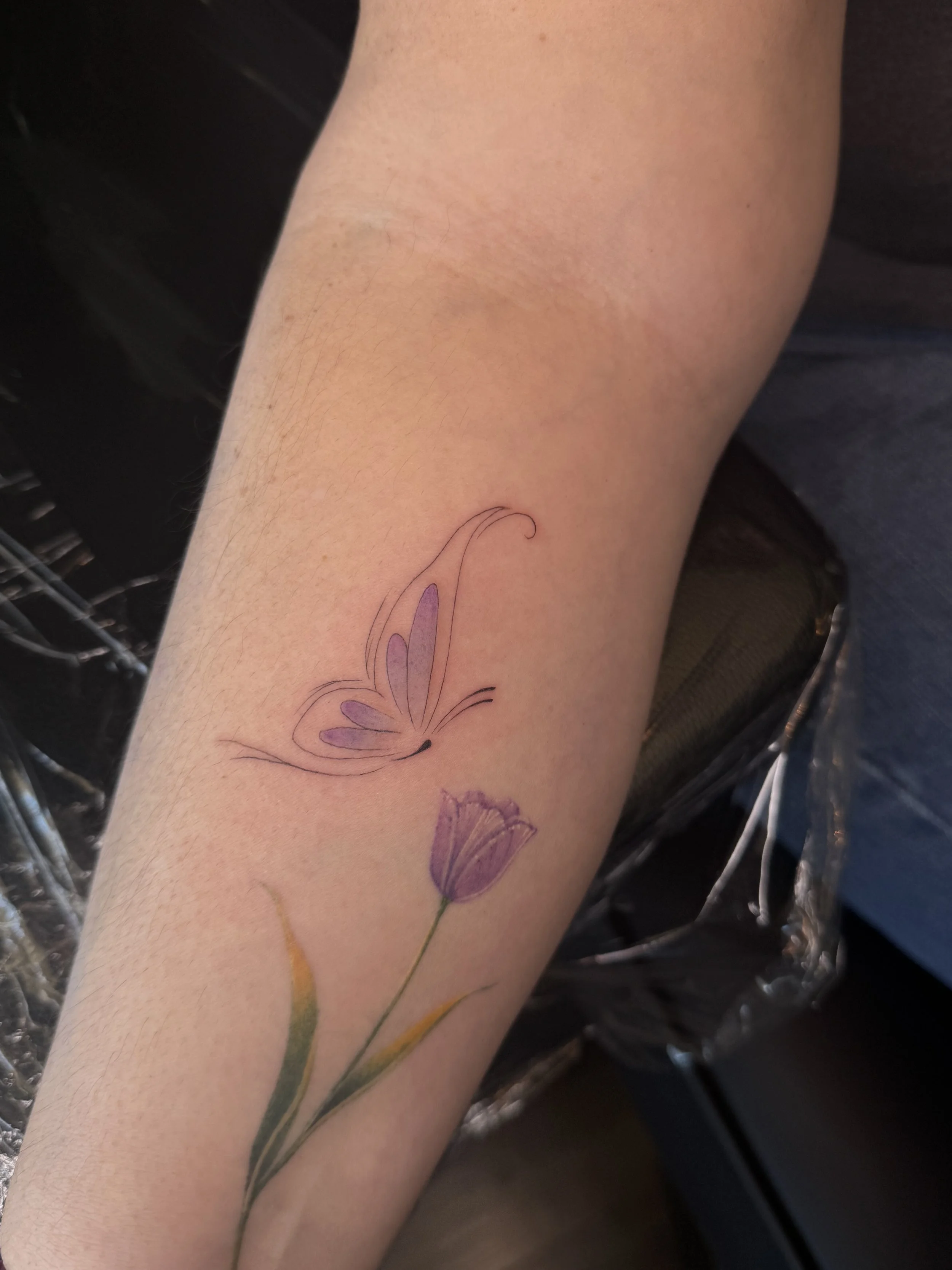 A person's arm with a delicate butterfly tattoo and a purple flower tattoo below it.