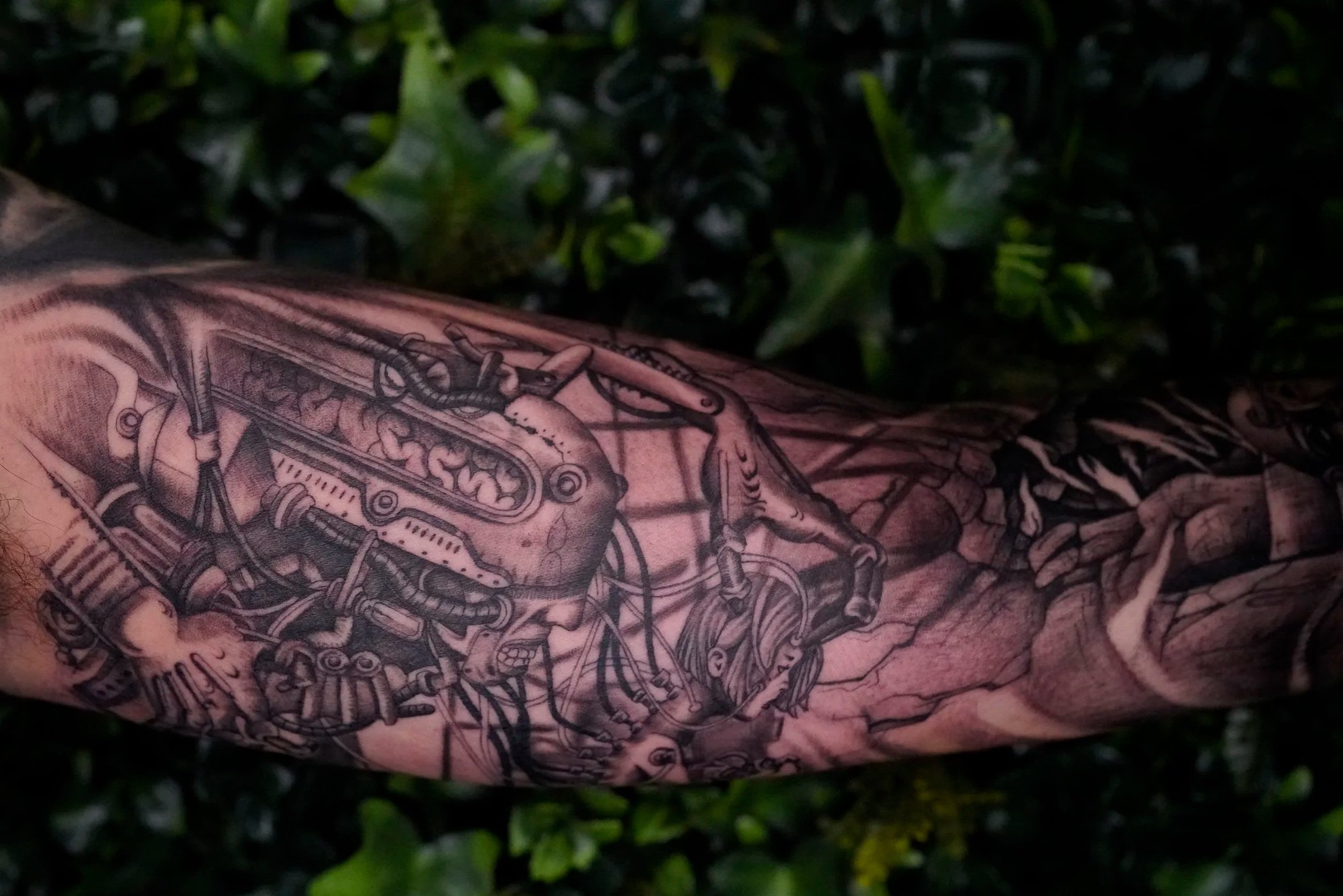 Tattooed arm with mechanical and anatomical designs, set against a leafy green background.
