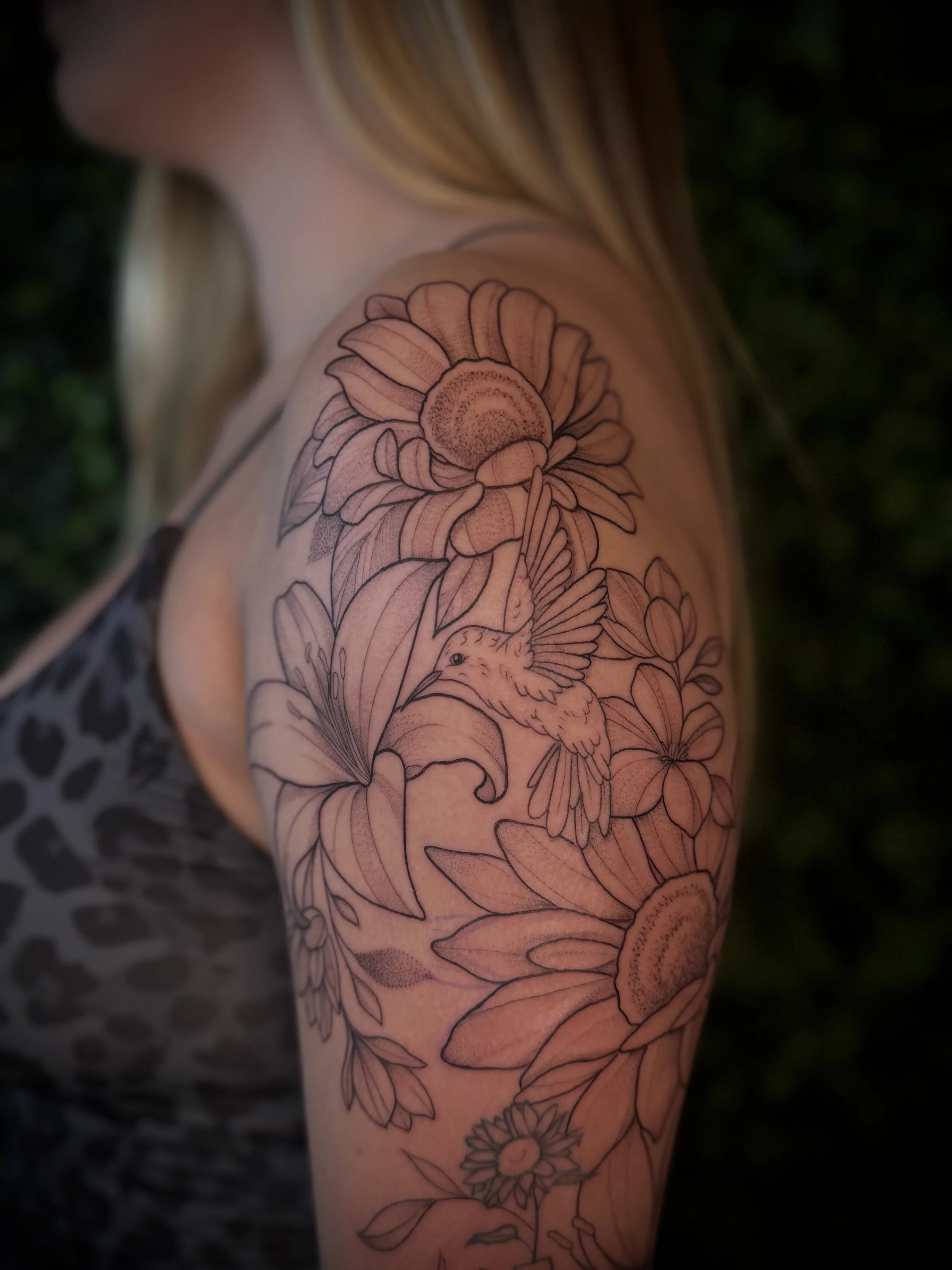 A person's arm with a floral tattoo featuring a coneflower and a hummingbird, with some greenery in the background.