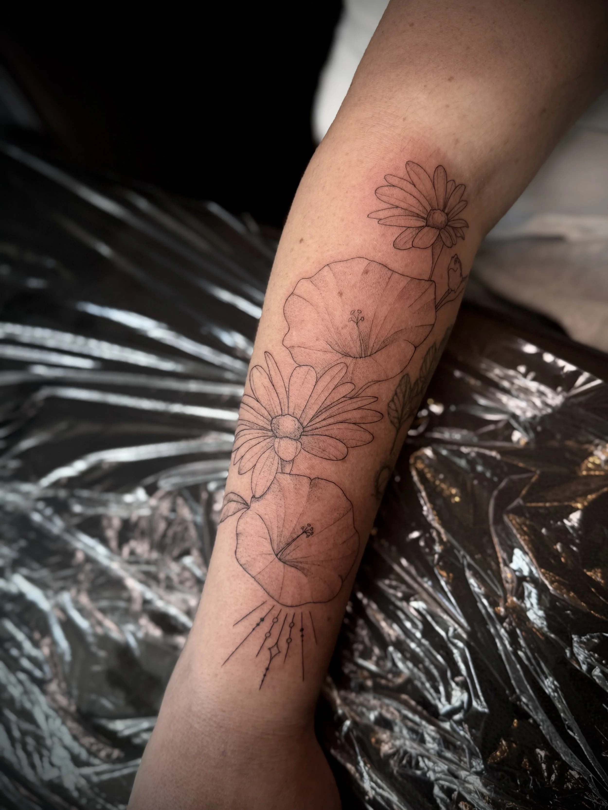 Line drawing of a daisy flower and a poppy flower tattooed on a person's forearm.