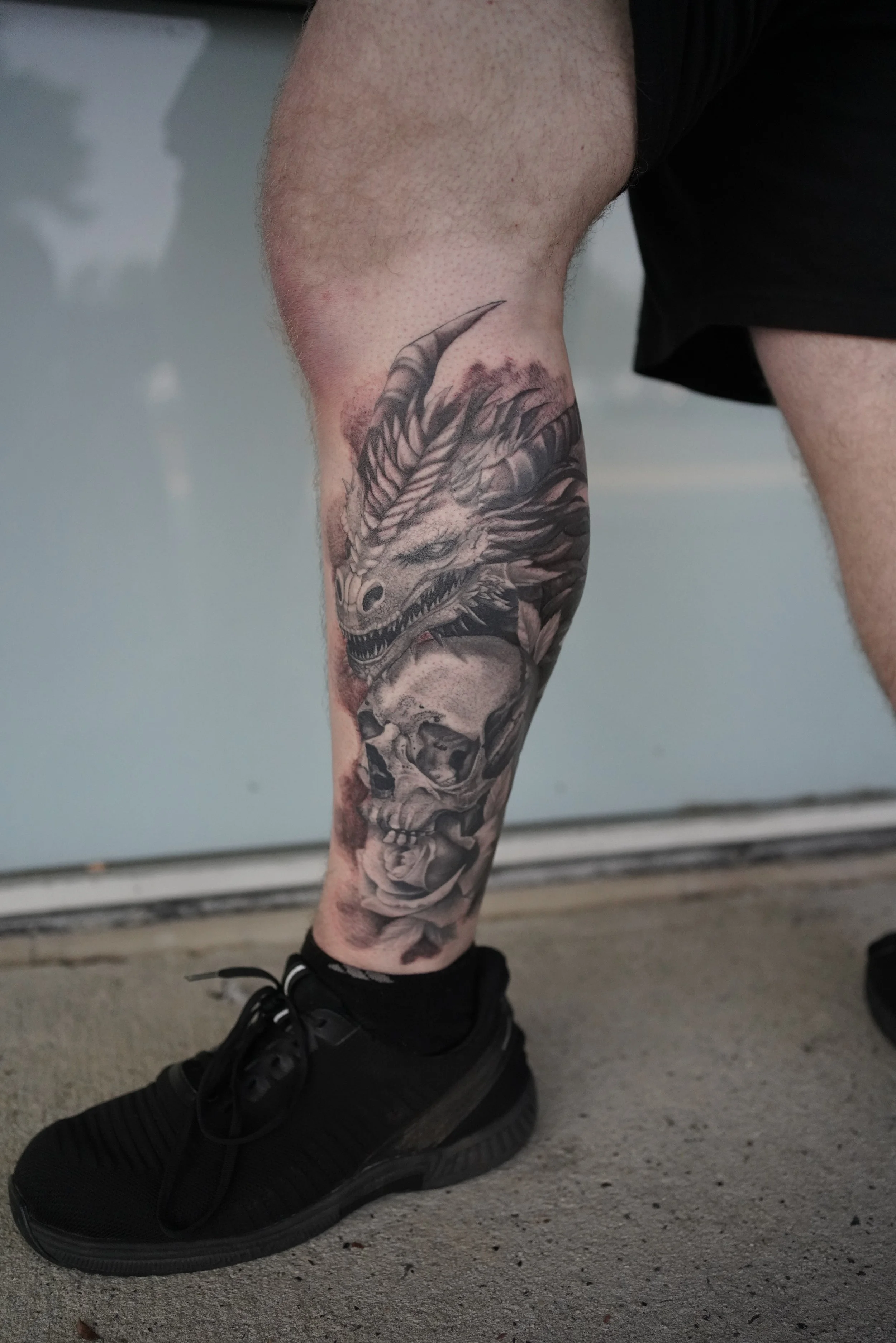 Tattoo of a dragon and a skull on a person's leg, with black sneakers and a black background.