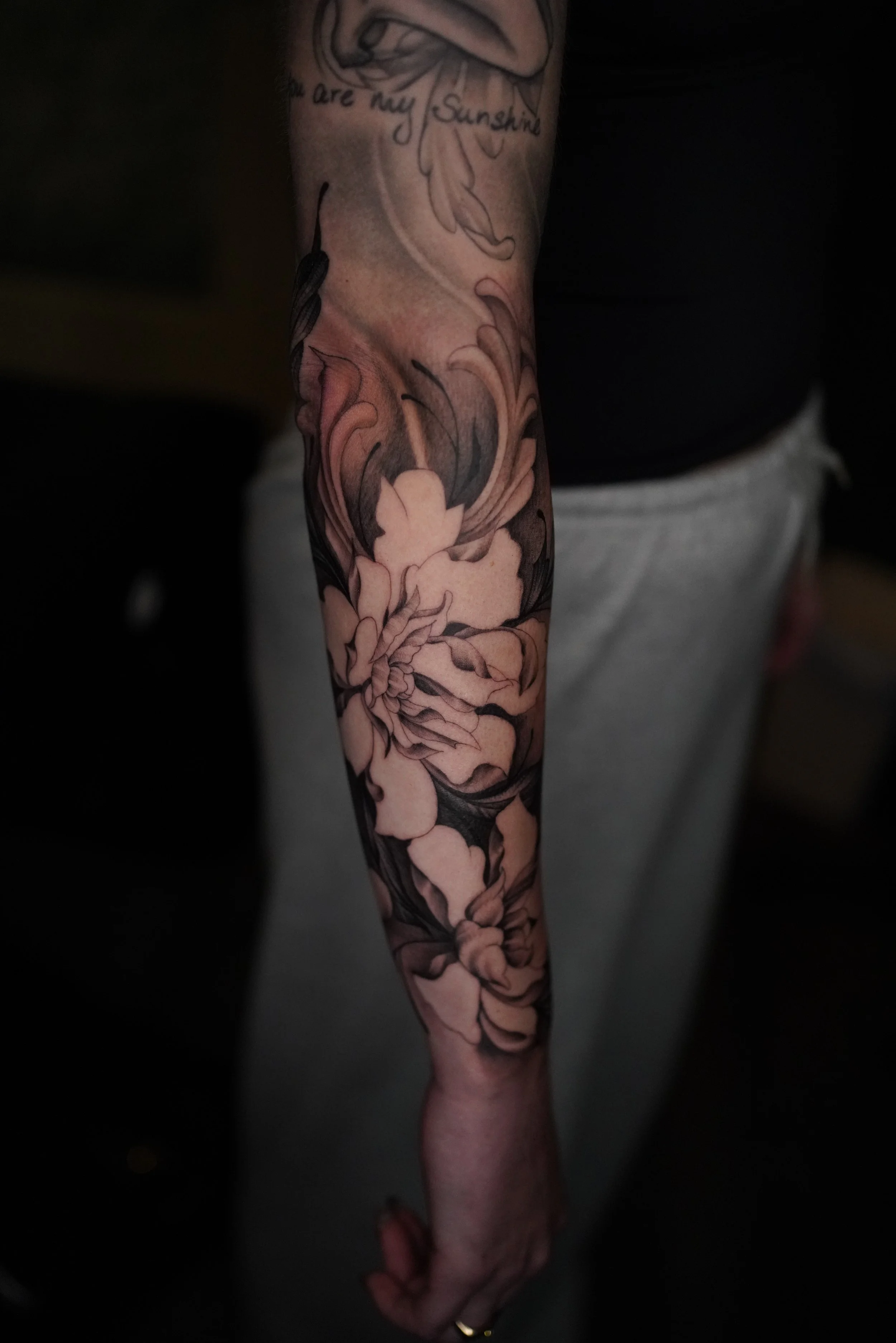 A person's arm with a large, detailed tattoo of flowers and leaves, with a partial quote that reads 'you are my Sunshine' near the top of the arm.