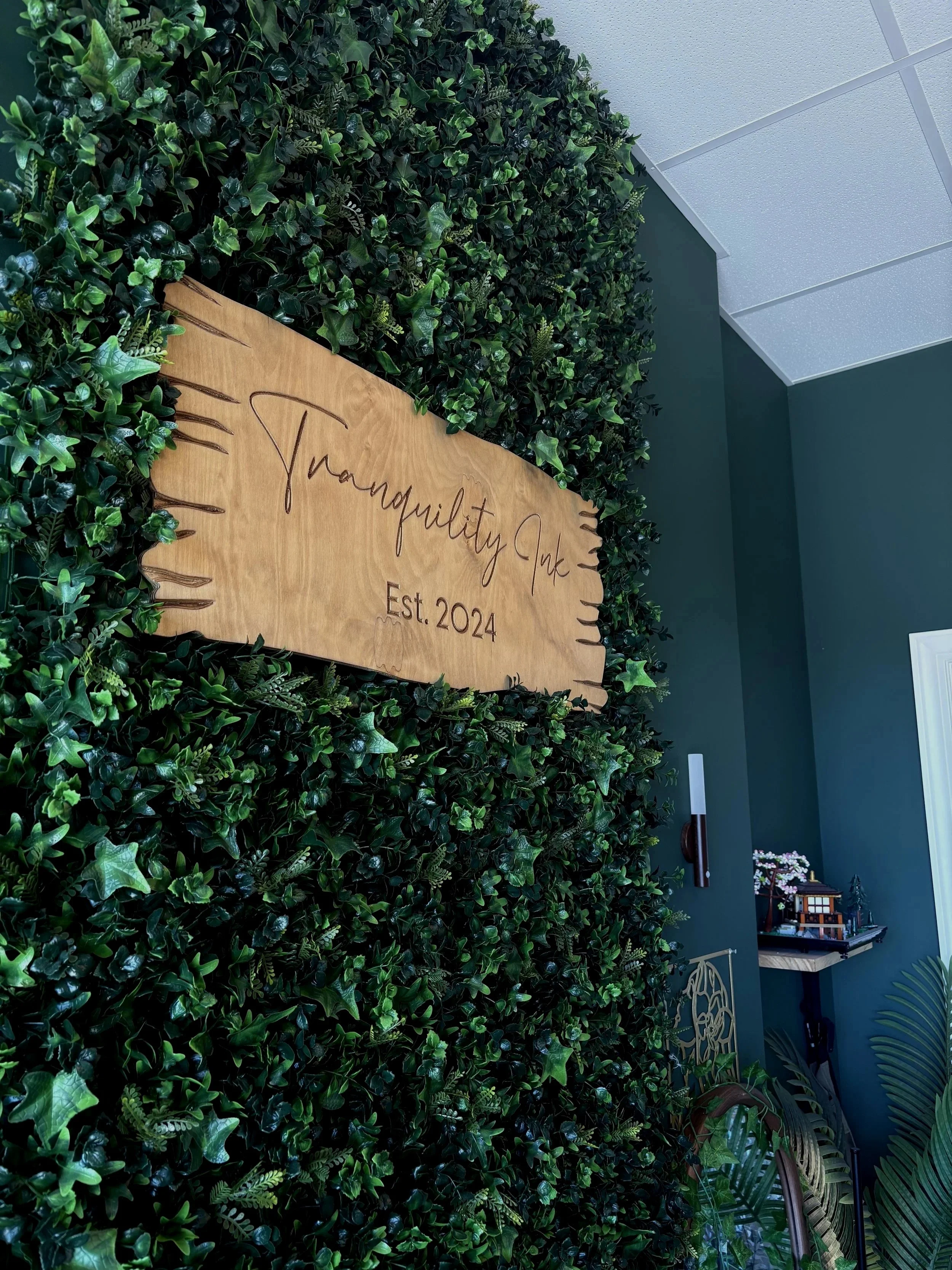 A wall covered with dense green ivy leaves with a wooden sign that reads 'Transquility Ink Est. 2024' mounted on it. The room has a green wall, ceiling tiles, and some decorative items in the background.