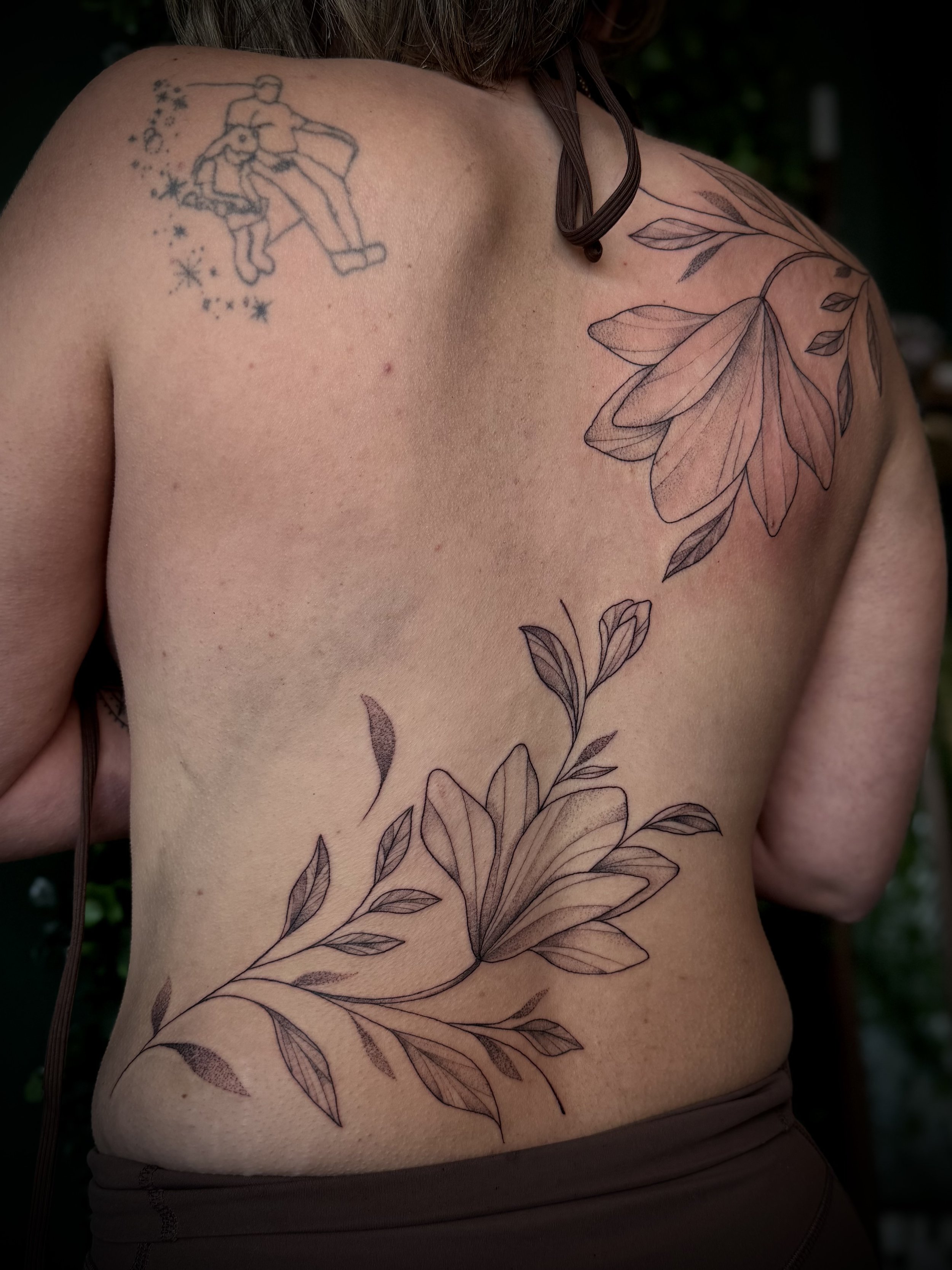 A person's shoulder and chest with several tattoos, including a delicate sketch of a large leaf or floral design on the chest and a small tattoo of a dancing couple surrounded by stars on the shoulder.