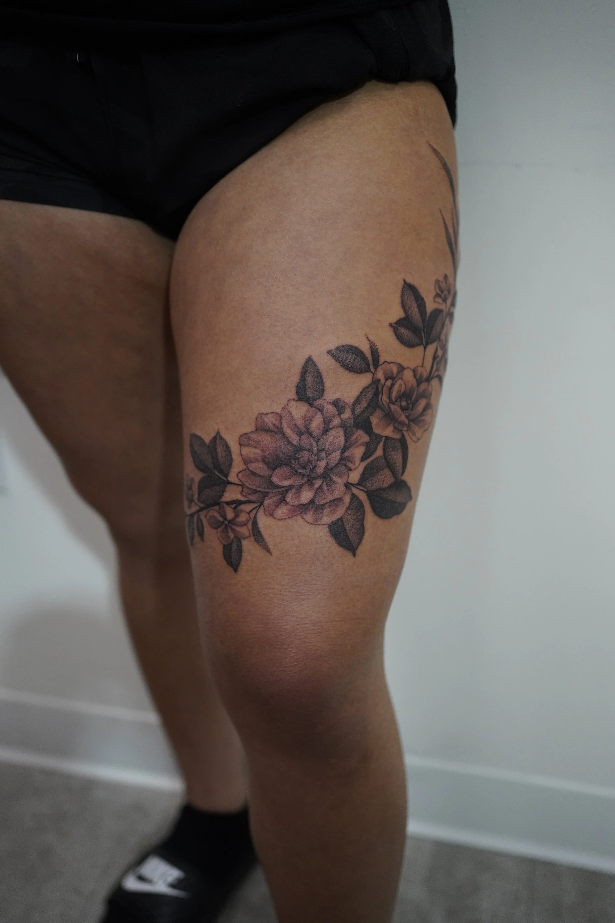 A person's thigh and knee showing a black floral tattoo on the thigh, with the person wearing black shorts and sneakers, standing indoors against a plain white wall.