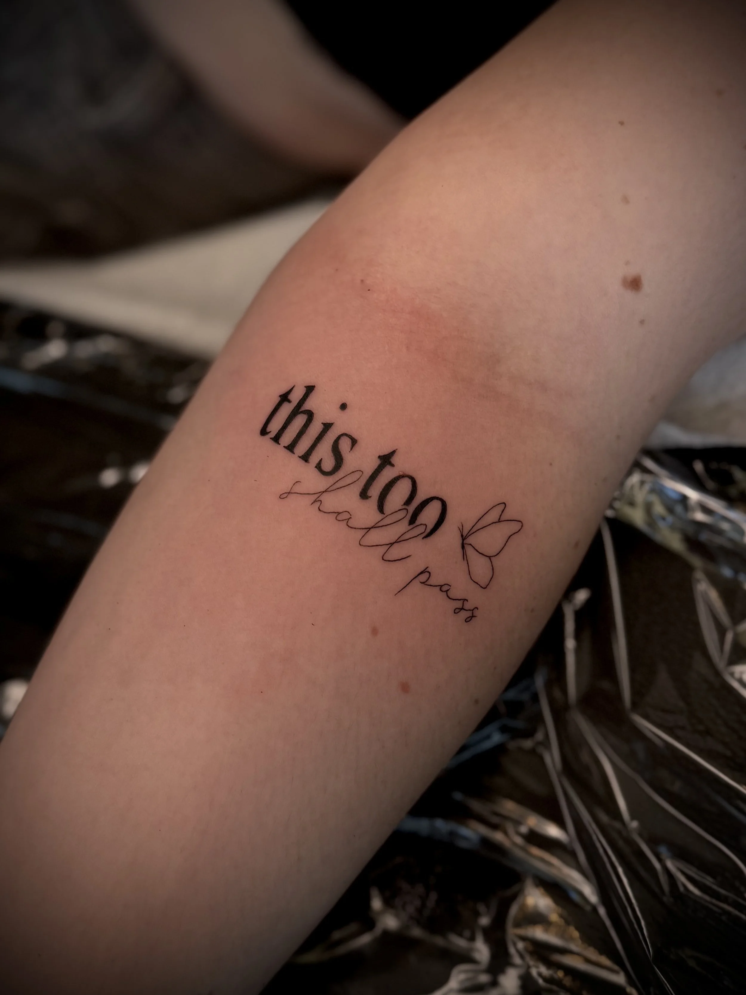 Close-up of a person's inner forearm with a tattoo that says "this too" and a small butterfly illustration underneath. The background includes dried or silver-colored plant stems.