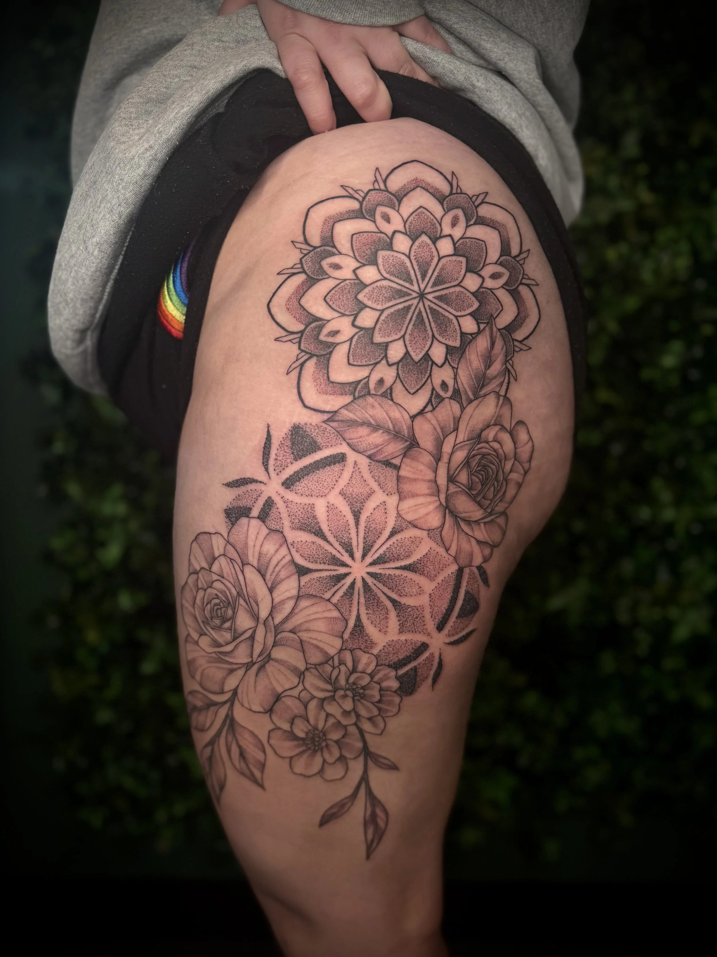 A person's thigh with a large black and gray tattoo of flowers, including roses, and decorative elements. The person is wearing gray and black clothing, and there is a rainbow rainbow patch on their clothing. They are outdoors with a green blurred ba