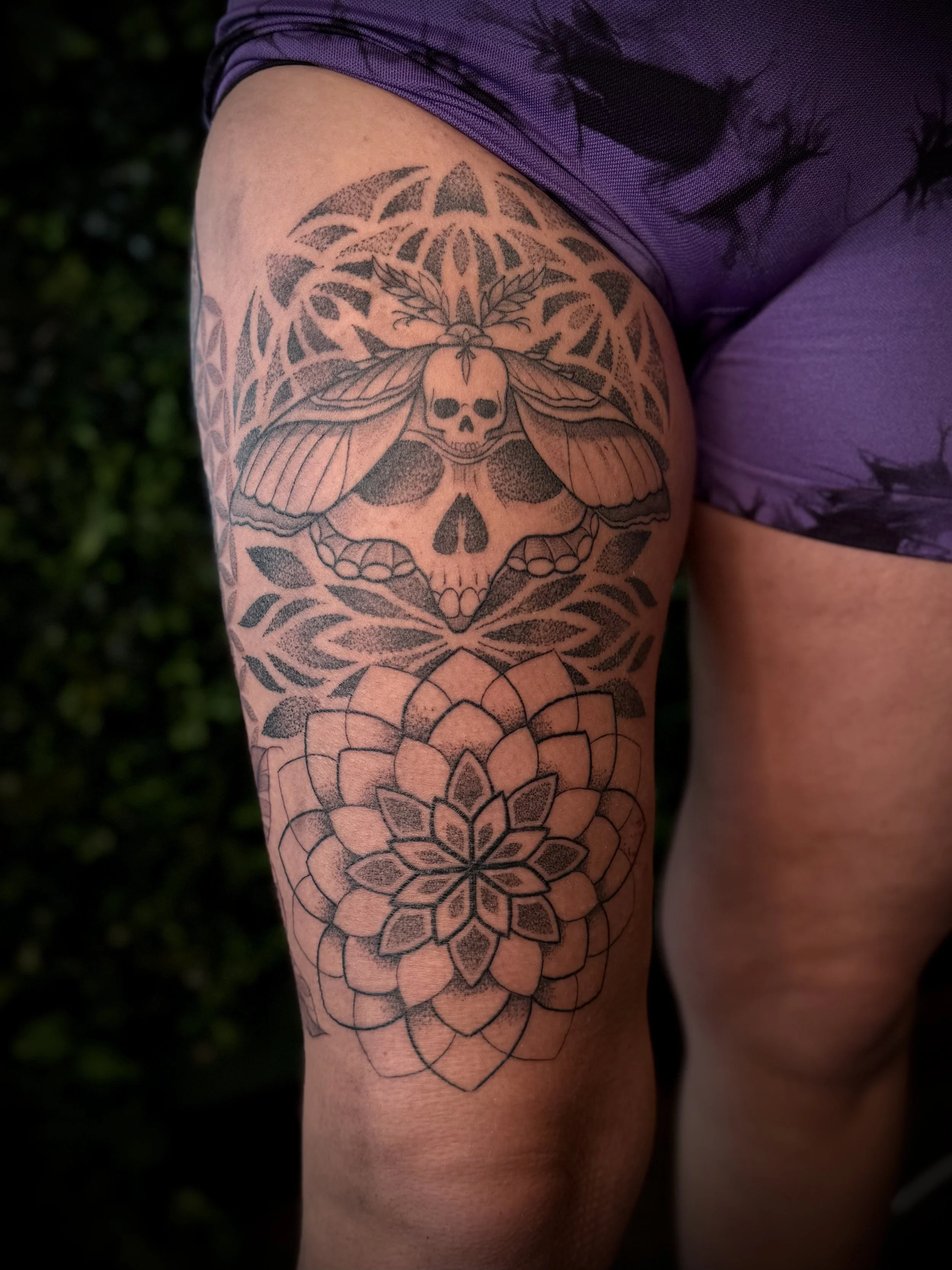Tattoo of a skull with moth wings and laurel leaves on the upper thigh, with a geometric and floral pattern below, against a background of a purple tie-dye shirt.