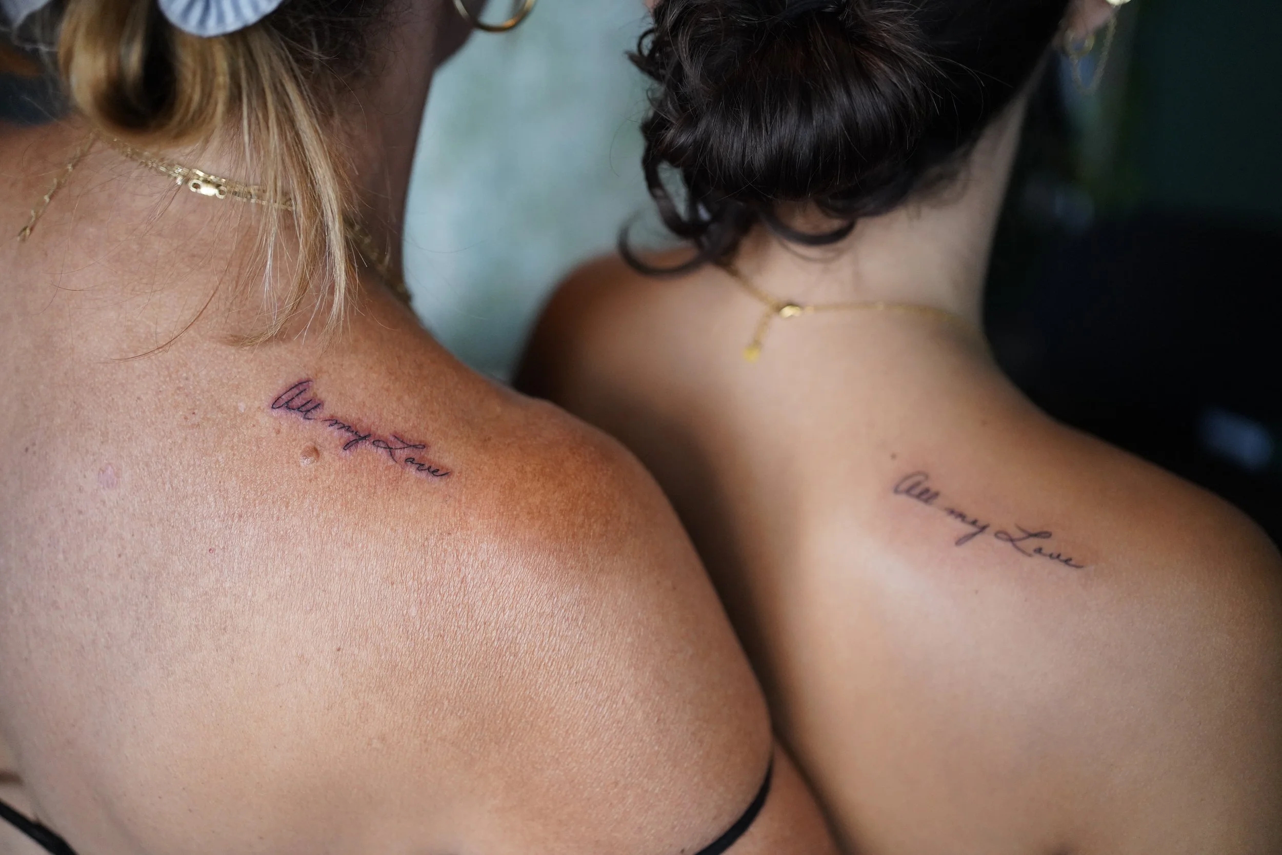 Close-up of two people's shoulders and necks, both with tattoos and wearing gold necklaces.