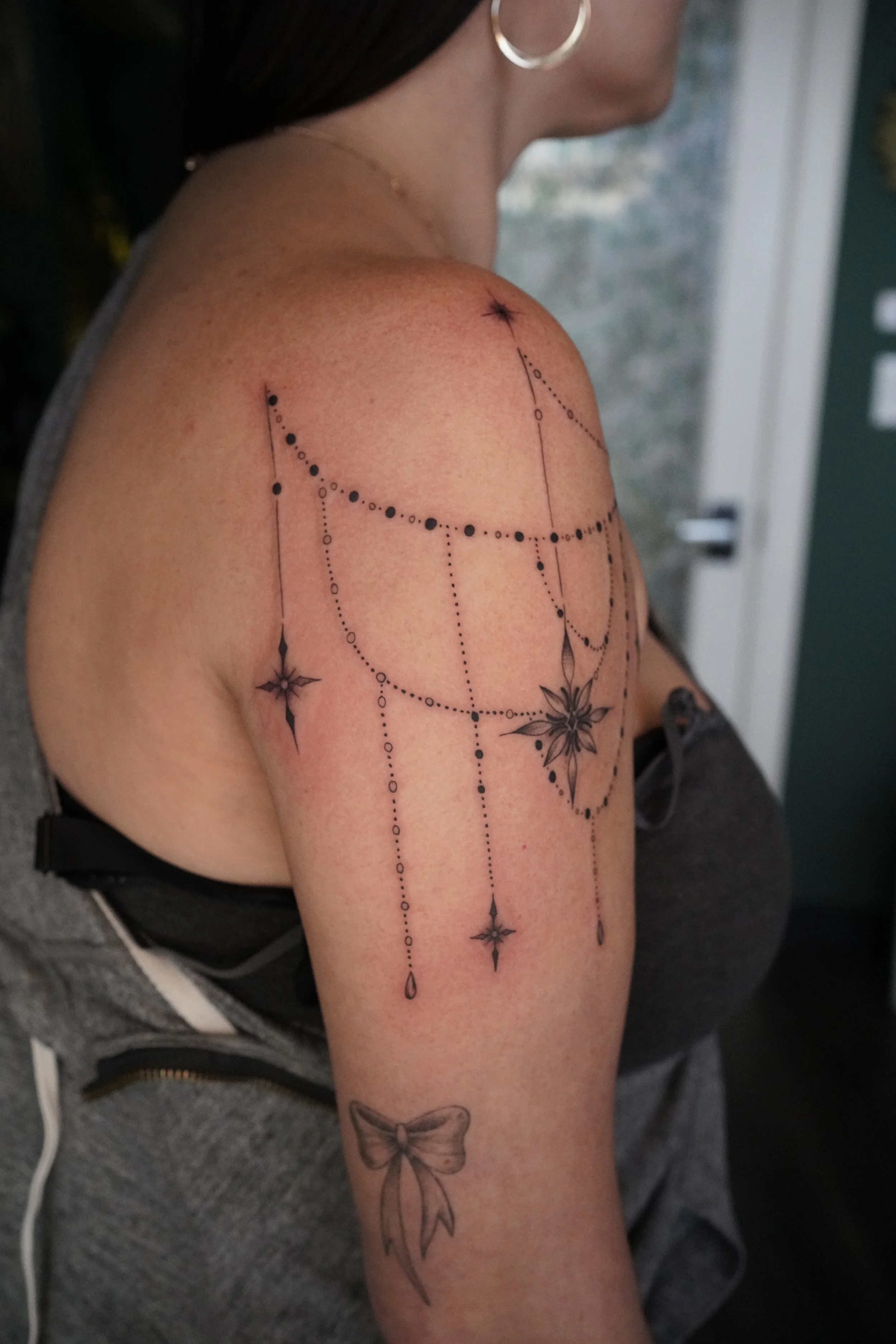 Close-up side view of a woman's upper arm and shoulder with a detailed black ink tattoo featuring a celestial theme with stars, dots, and a bow.