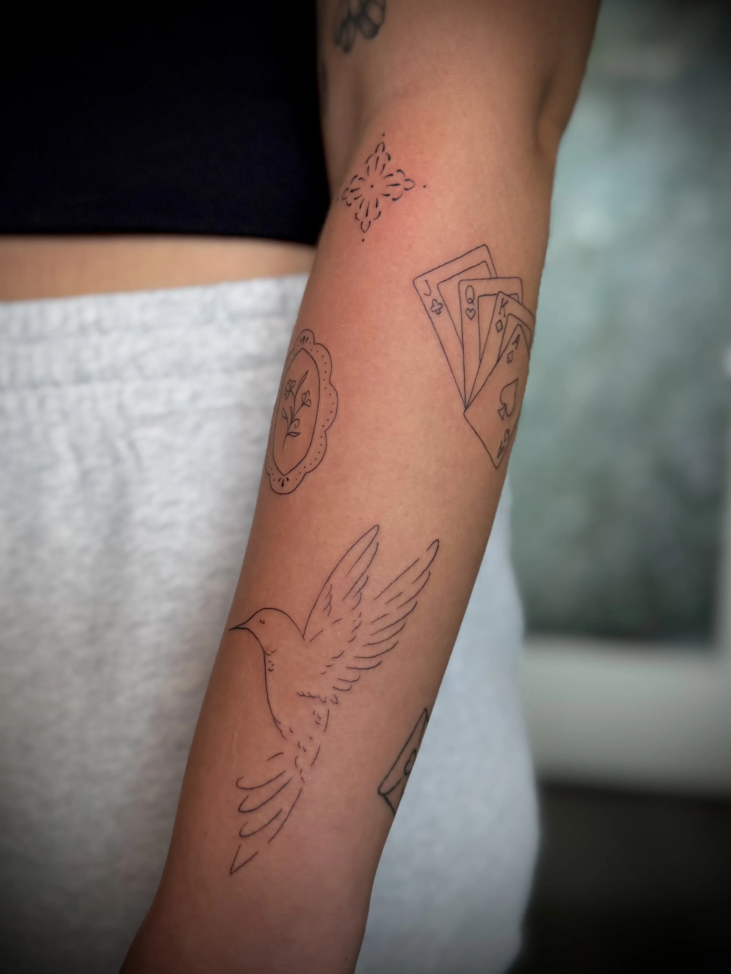 A person's arm with several minimalist line tattoos, including a dove, a pocket watch with flowers, and playing cards showing a royal flush of spades, hearts, diamonds, and clubs.