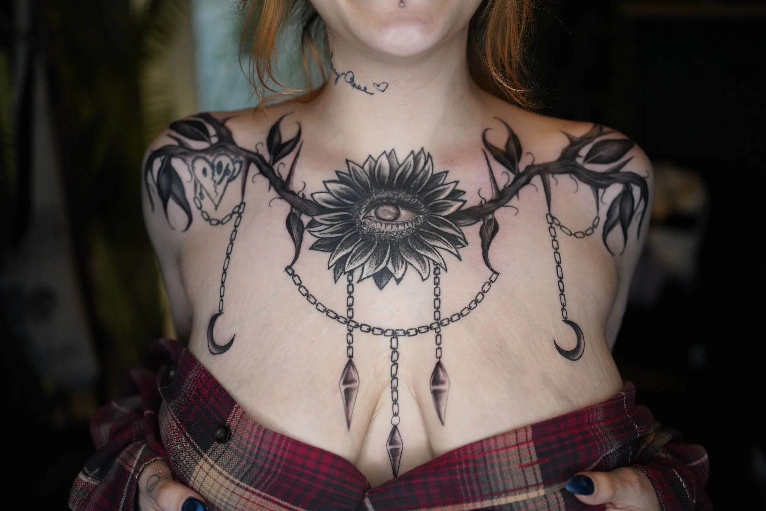 Close-up of a woman's chest and neck tattoos, featuring a sunflower with an eye in the center, hanging chains with moon-shaped charms, and a small handwritten tattoo that reads 'love' with a heart. She is wearing a red plaid shirt.