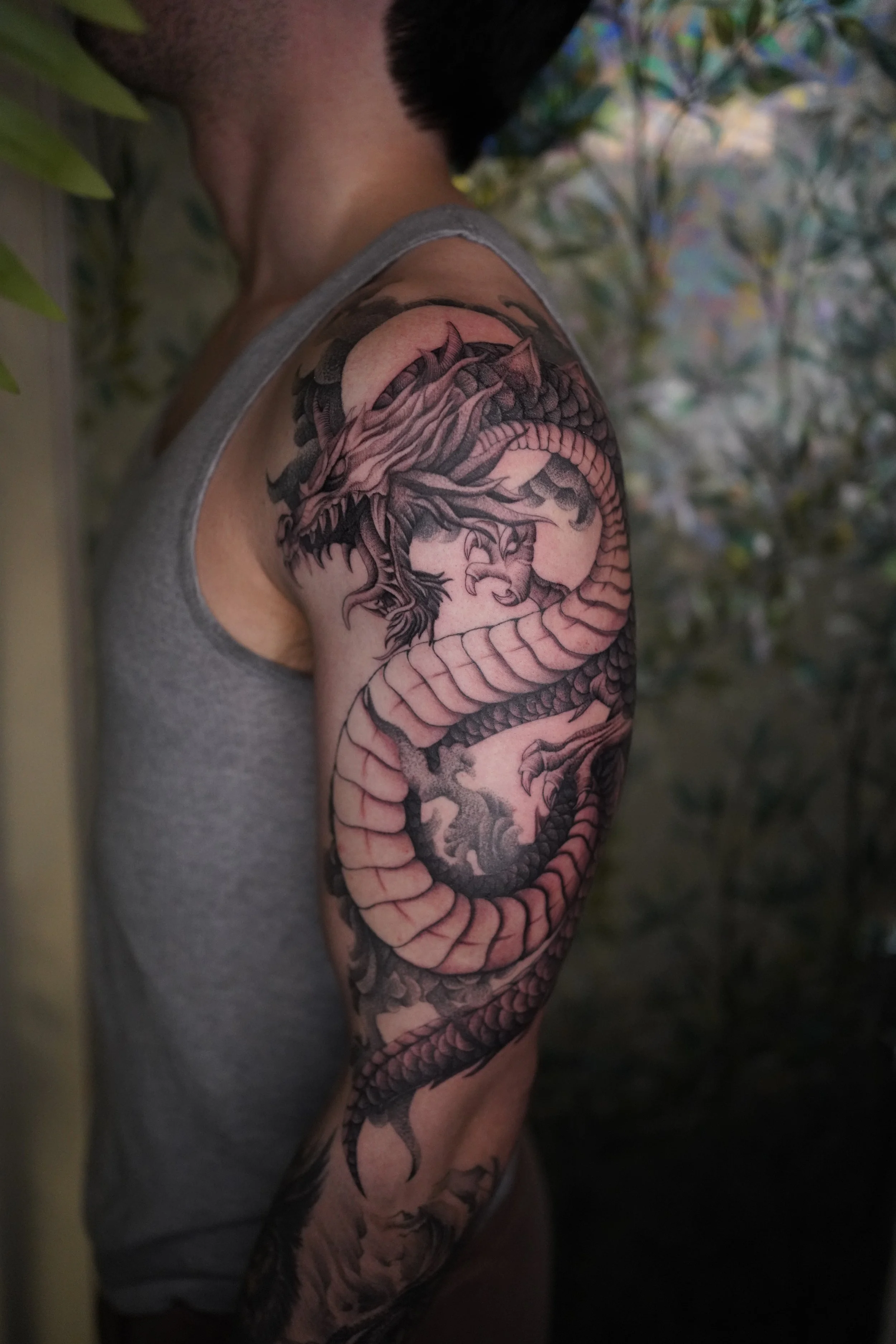 Close-up view of a person's upper arm showing a detailed black and gray tattoo of a dragon wrapping around the arm, with scales and claws visible, and a background of leaves and blurred colorful foliage.