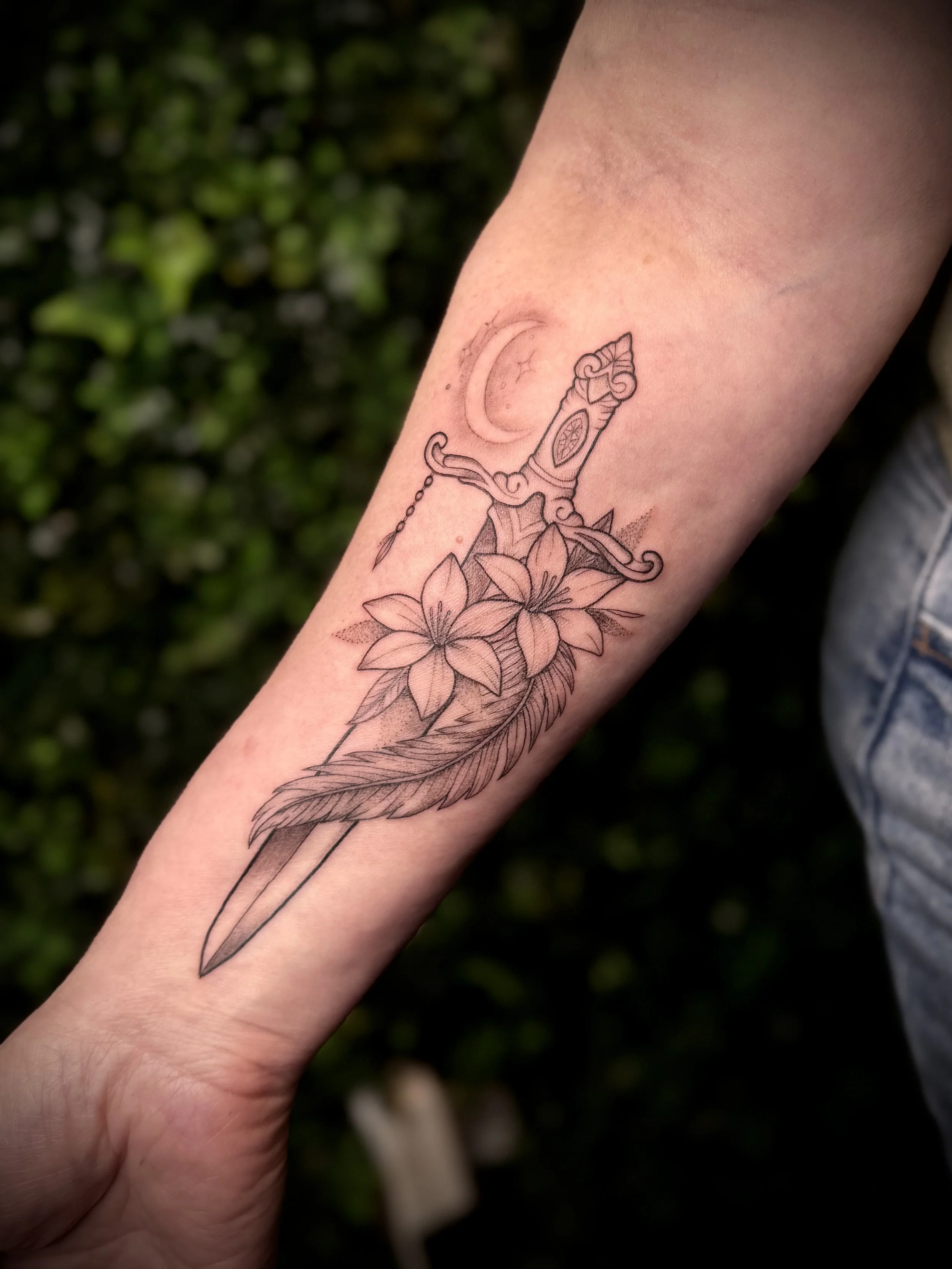 Tattoo of a dagger piercing a flower, with a crescent moon in the background on person's forearm.