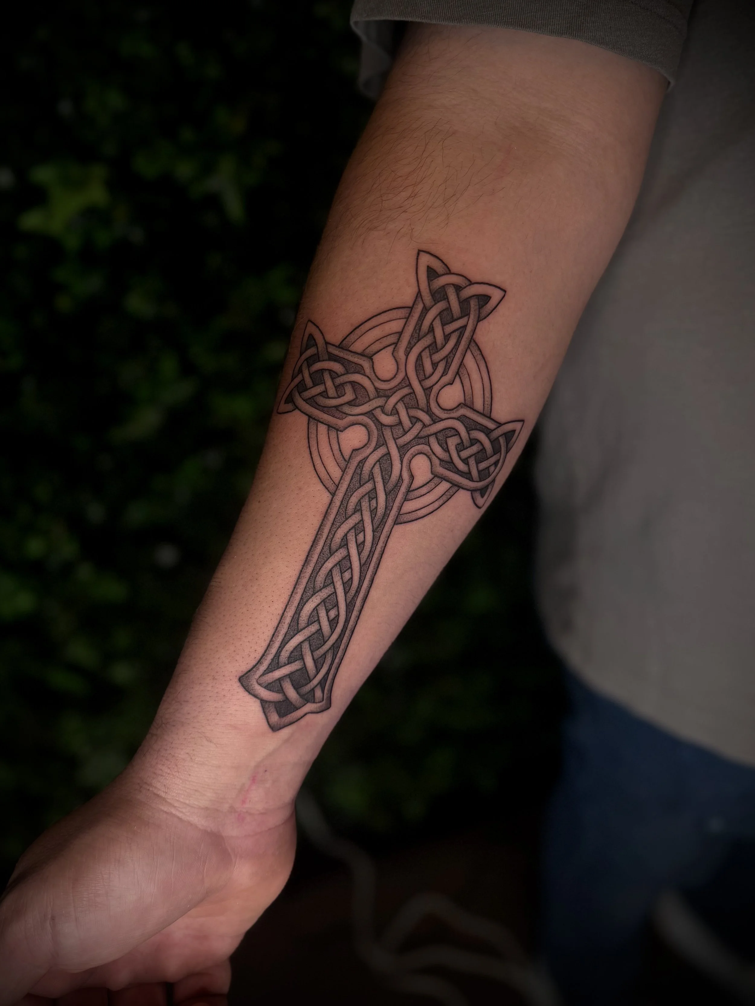 Close-up of an arm with a detailed black and gray Celtic knot tattoo on the inner forearm, with a blurred background of green foliage.