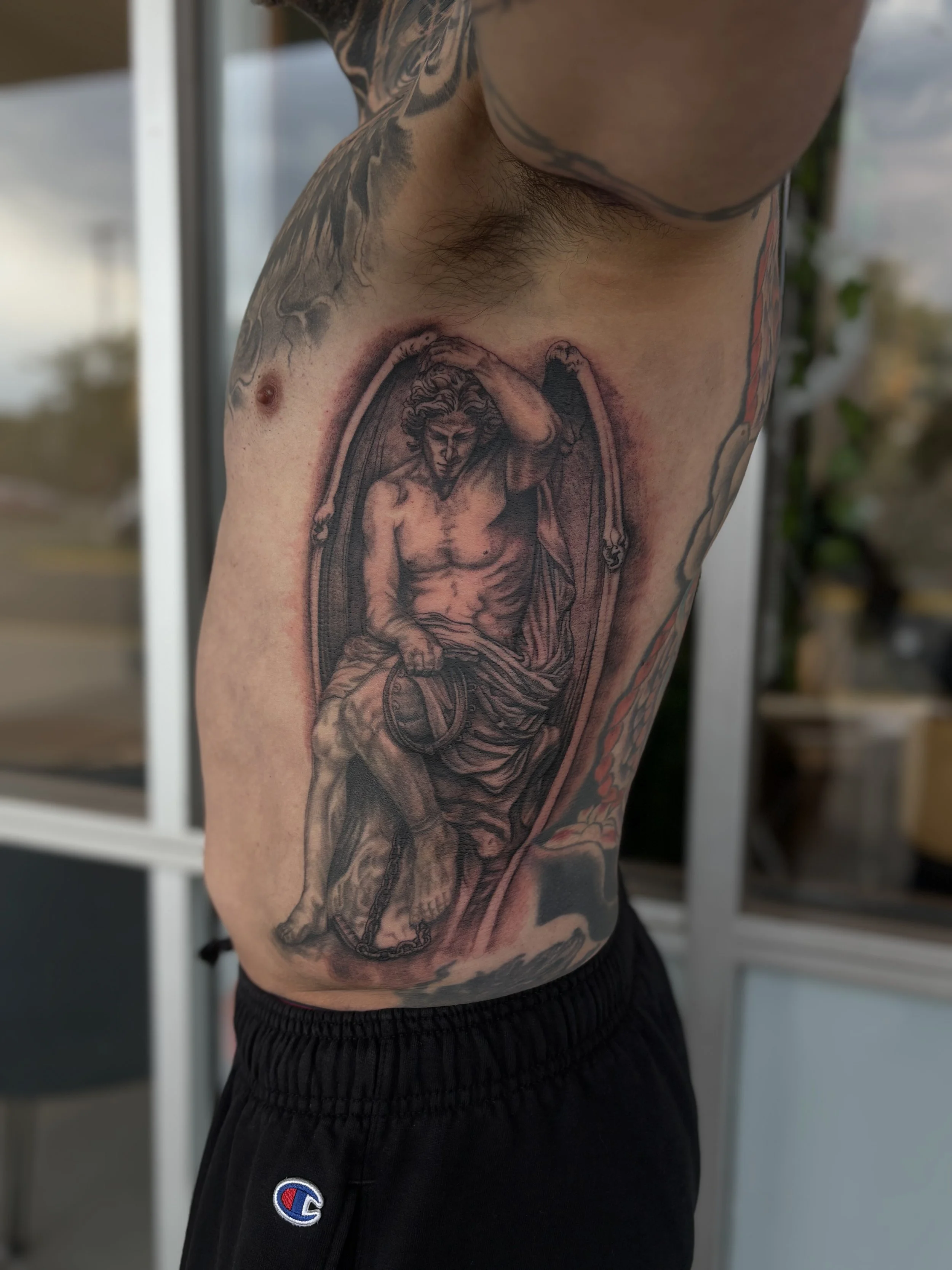 Close-up of a person's side torso showing a tattoo of an angel holding a shield, with additional tattoos on the arm and chest, and wearing black Champion shorts.