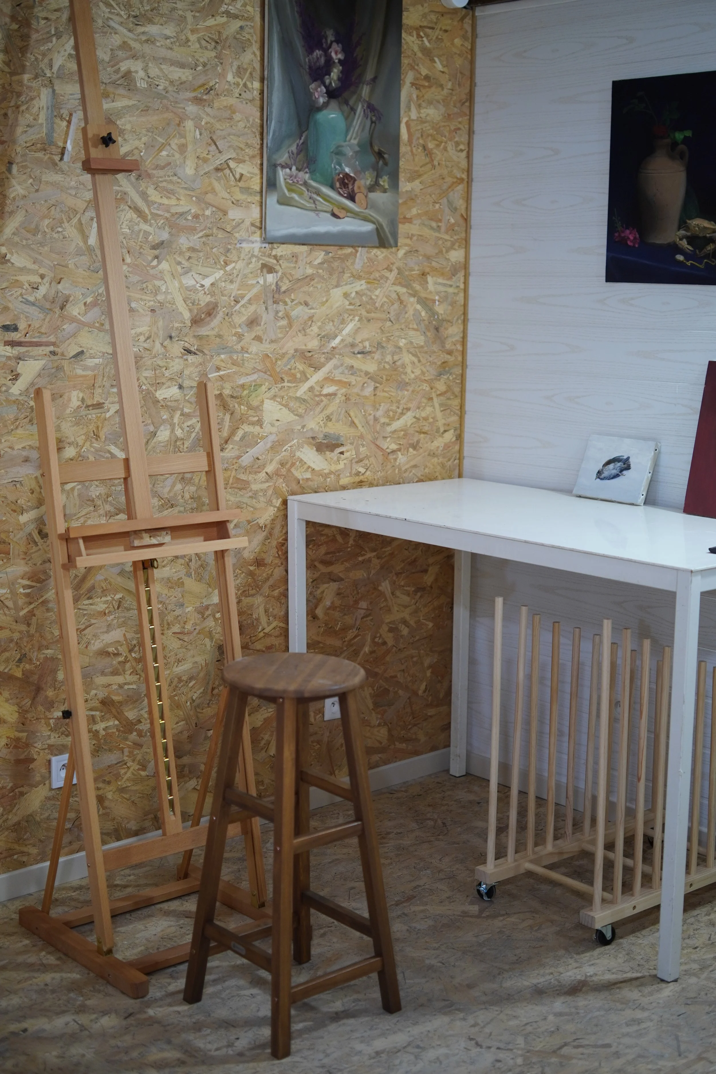 Interior of an art studio with plywood wall and white table, art supplies, and paintings on the wall.