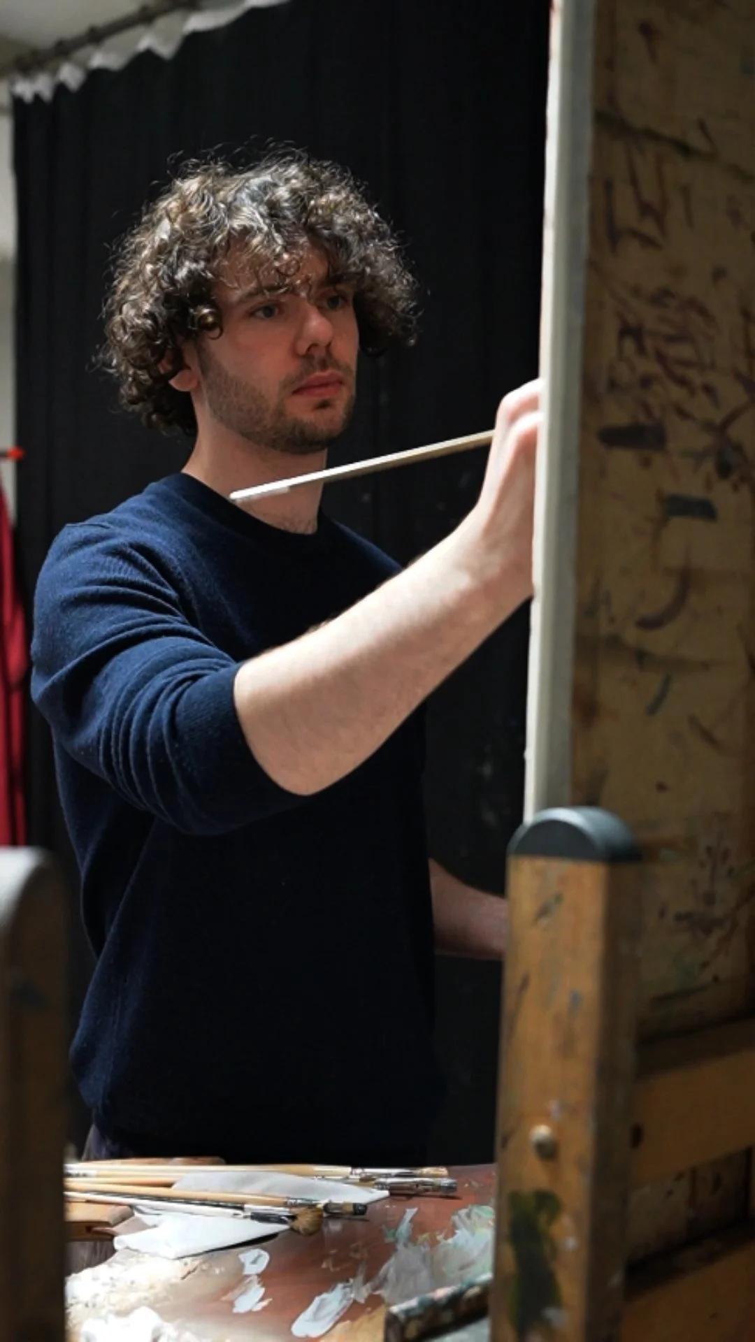A man with curly dark hair and a beard standing in front of an easel, painting with a brush during an art session.