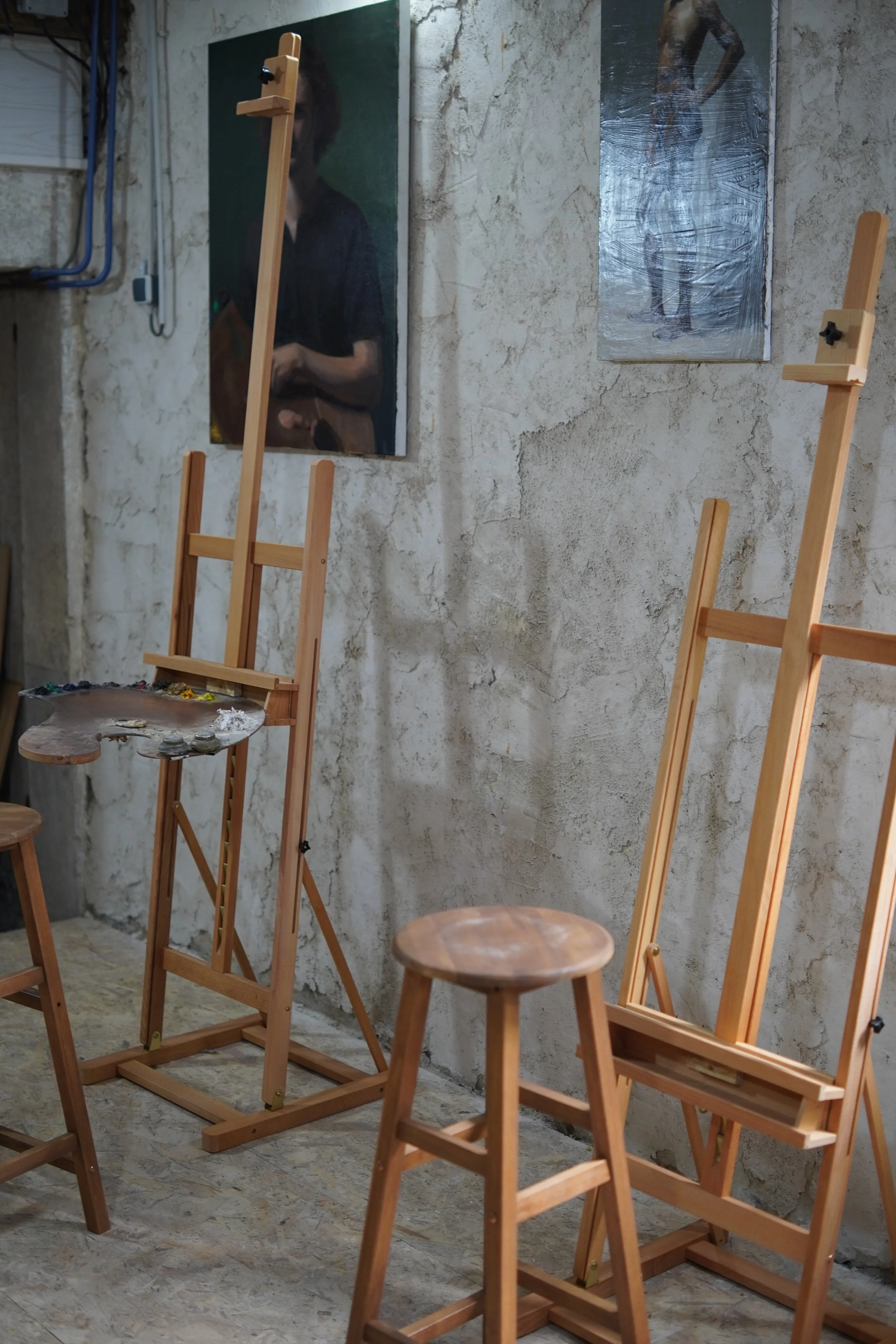 A cozy art studio with unfinished wooden easels, a painting palette, and two paintings on a textured concrete wall.