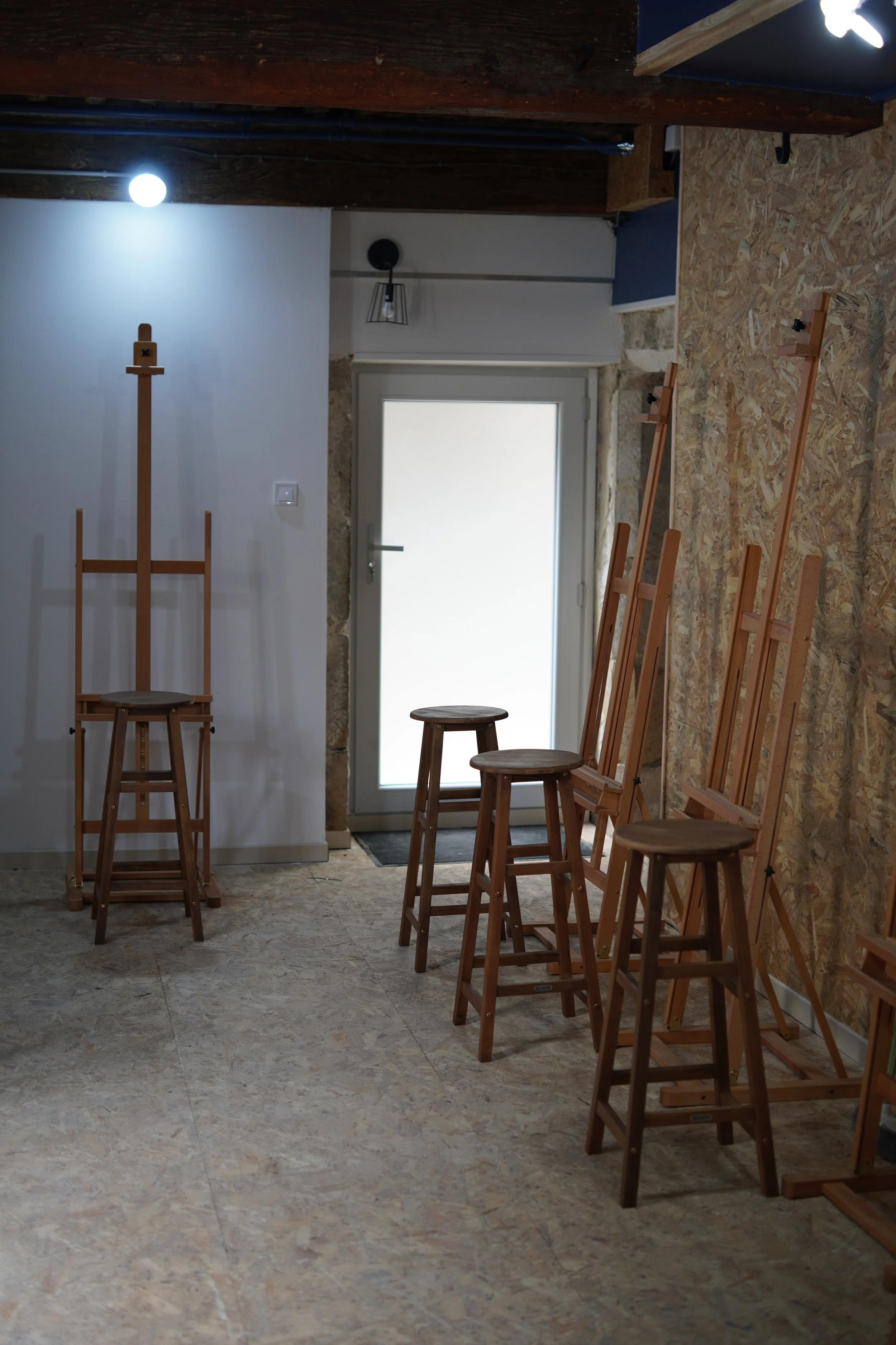 Interior of a room with wooden stools, some on the floor and some leaning against the wall, and an unfinished wall with a wooden frame. There is a door with frosted glass and a stone wall surrounding it. The floor is made of light-colored tiles.