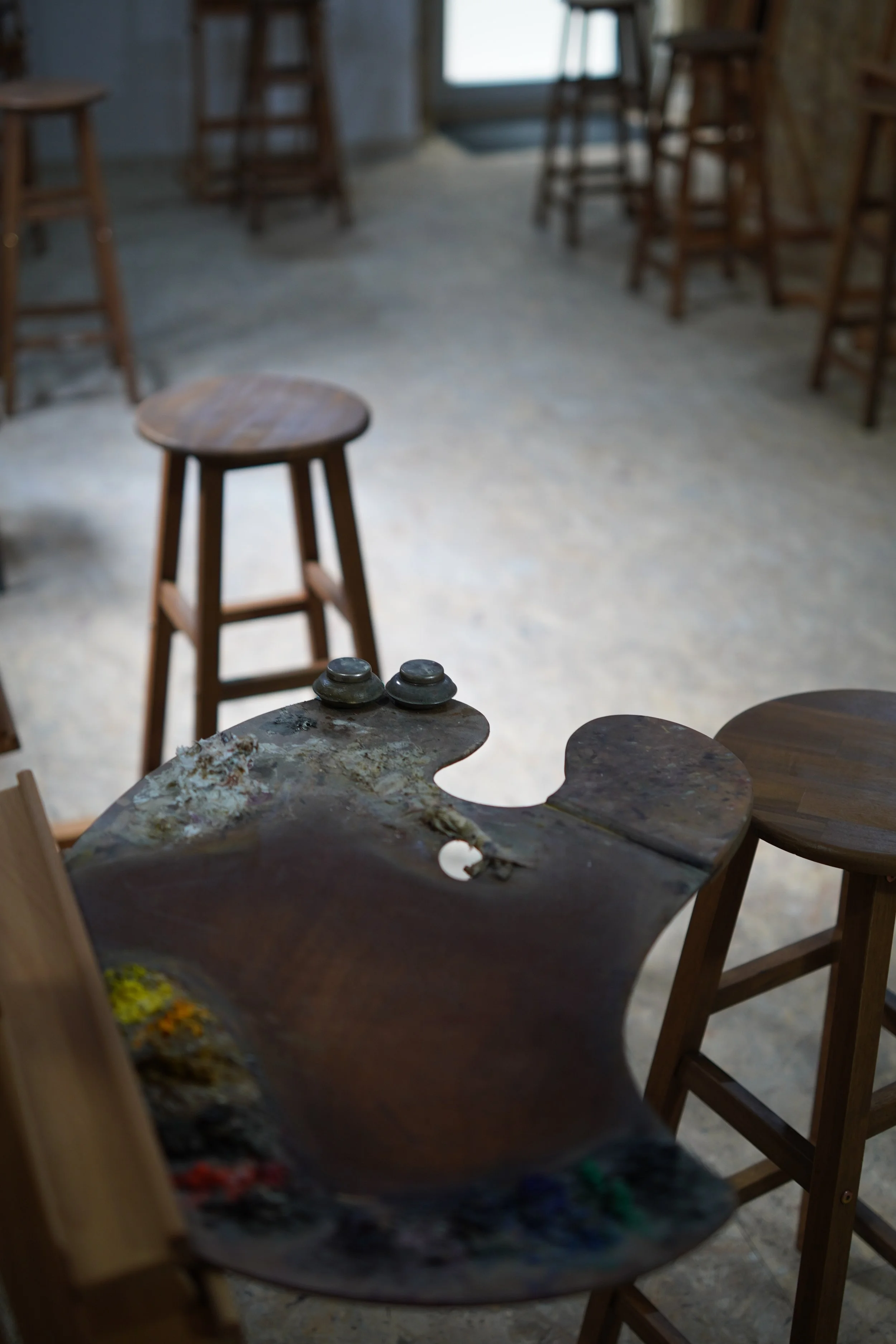 Wooden stools and a table with paint splatters in an art studio or classroom setting with natural light coming from the window.