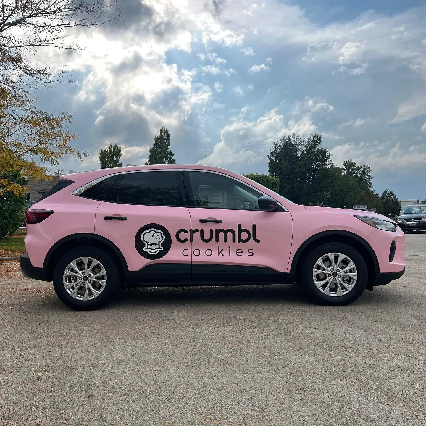 Pink SUV with Crumbl Cookies logo and text on the side, parked outdoors under a cloudy sky.