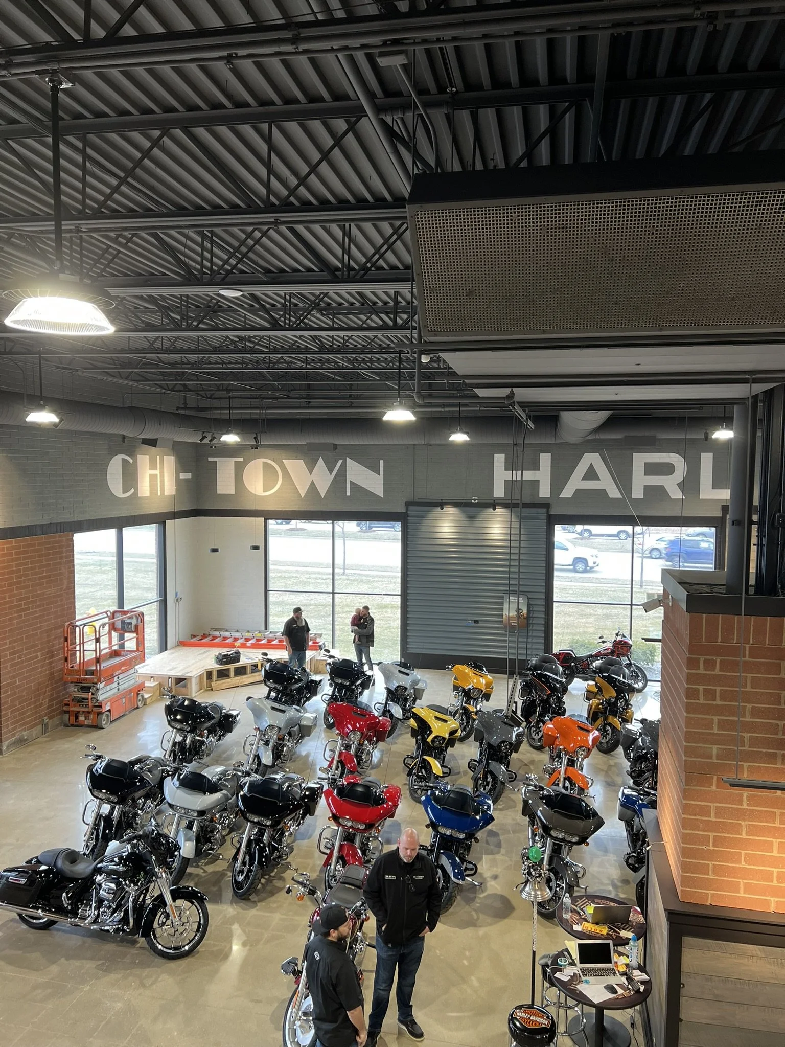 Indoor motorcycle showroom with multiple motorcycles displayed in various colors. Several people are present, including a man in a Harley Davidson jacket. The space has large windows, brick accents, and a high, industrial-style ceiling with exposed m