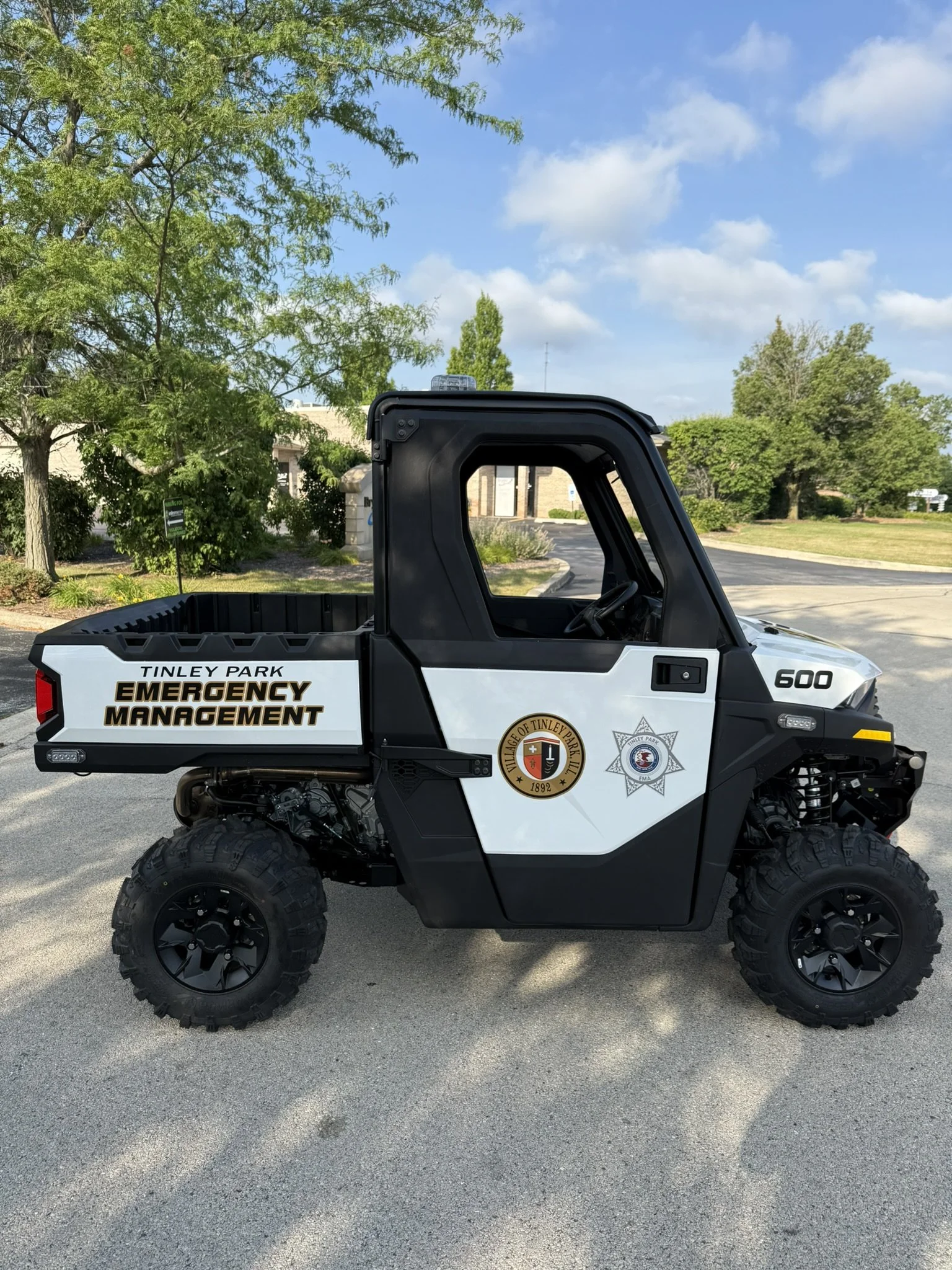 Off-road utility vehicle with 'Tinley Park Emergency Management' logo, black and white color scheme, parked on pavement with green trees and blue sky in the background.