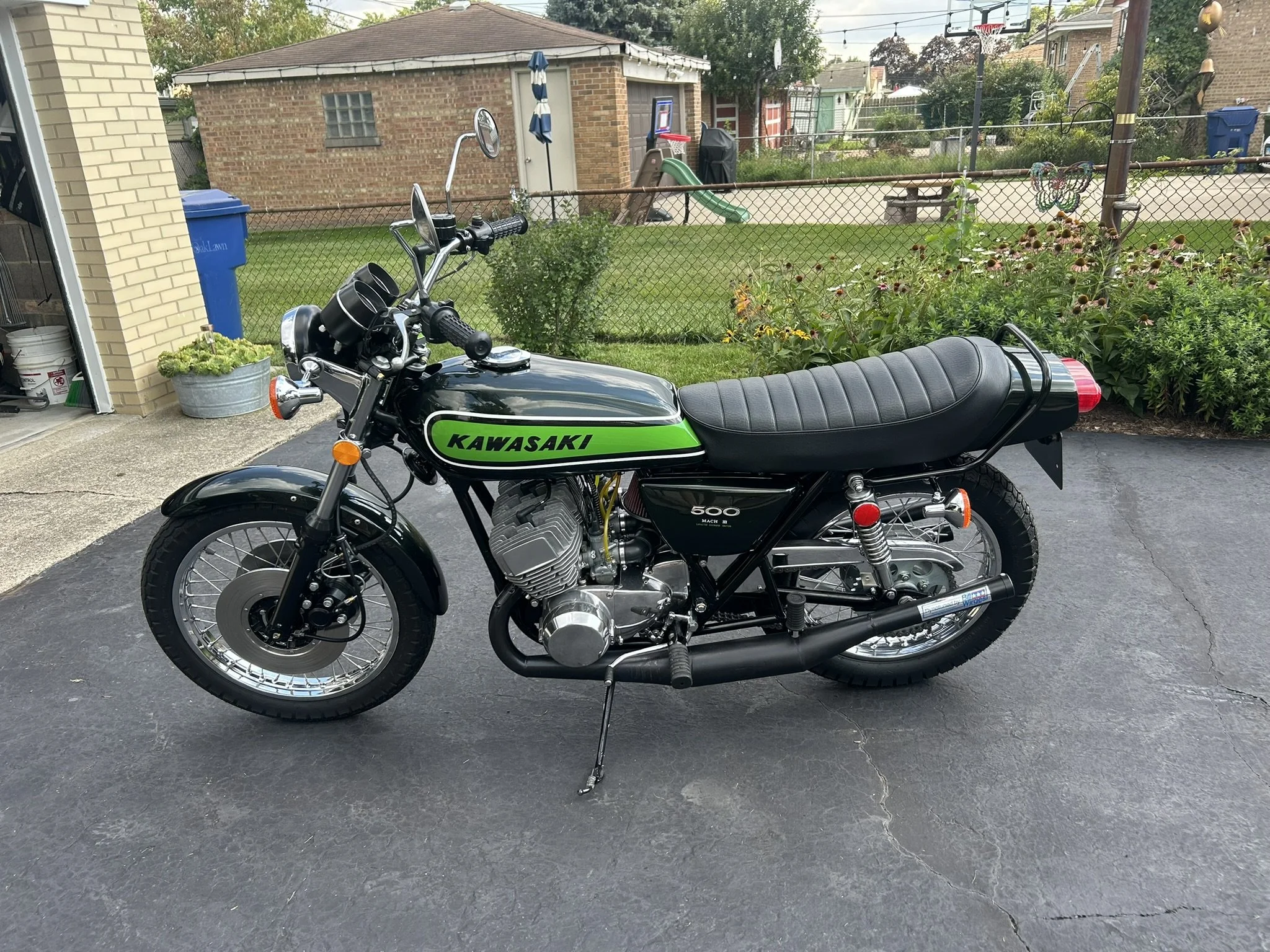 A vintage black Kawasaki motorcycle with a green and black logo, parked on a driveway in front of a backyard. The backyard has a chain-link fence, a small playset with a slide, and various plants and flowers.
