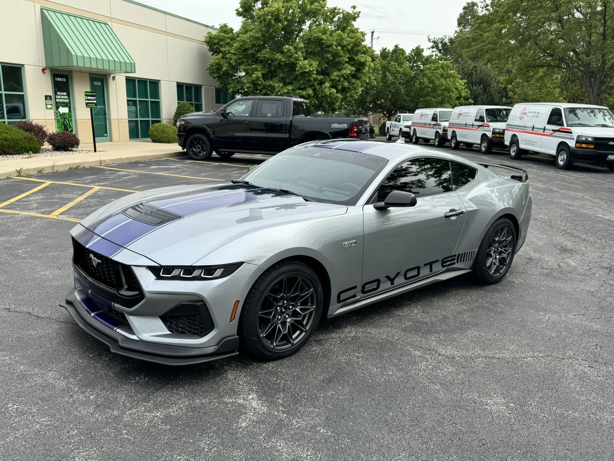 A silver sports car with a black stripe and the word 'GOTYOTE' on its side parked in a parking lot in front of a building with green awnings. Behind it, a black pickup truck and several white vans with red and black stripes are parked. Green trees su