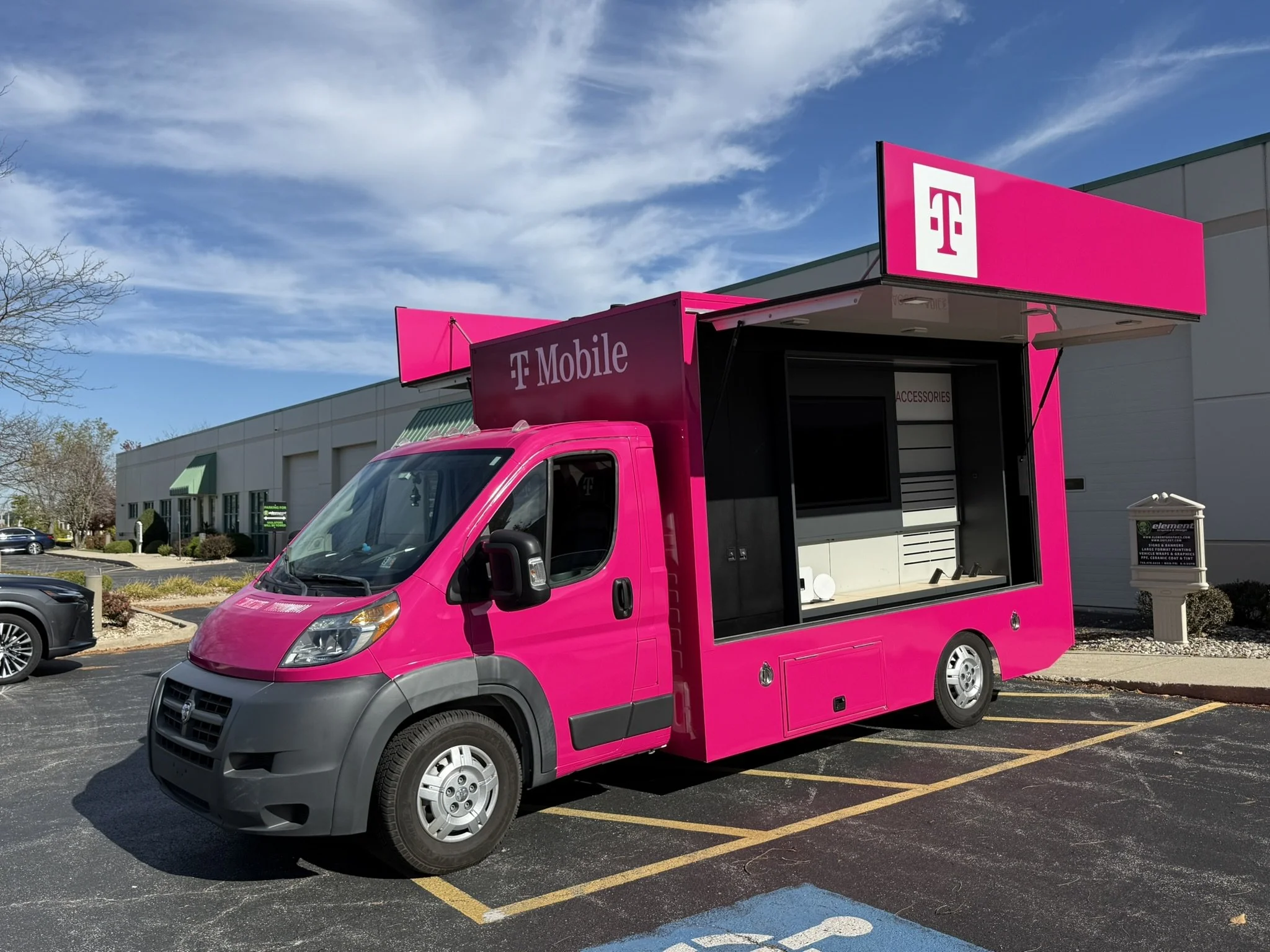 A pink mobile store truck with T-Mobile branding parked in a parking lot on a sunny day. The truck has an open window showcasing accessories inside.