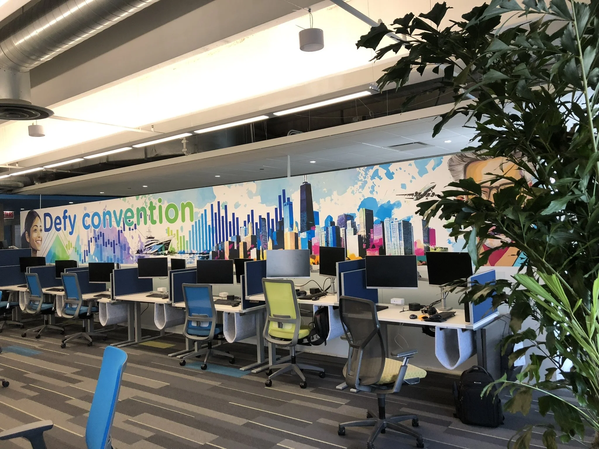 An office space with multiple workstations arranged in a row, each equipped with computers and monitors. In the background, there is a colorful mural depicting a city skyline with the text "Defy convention." A large indoor plant partially obscures th