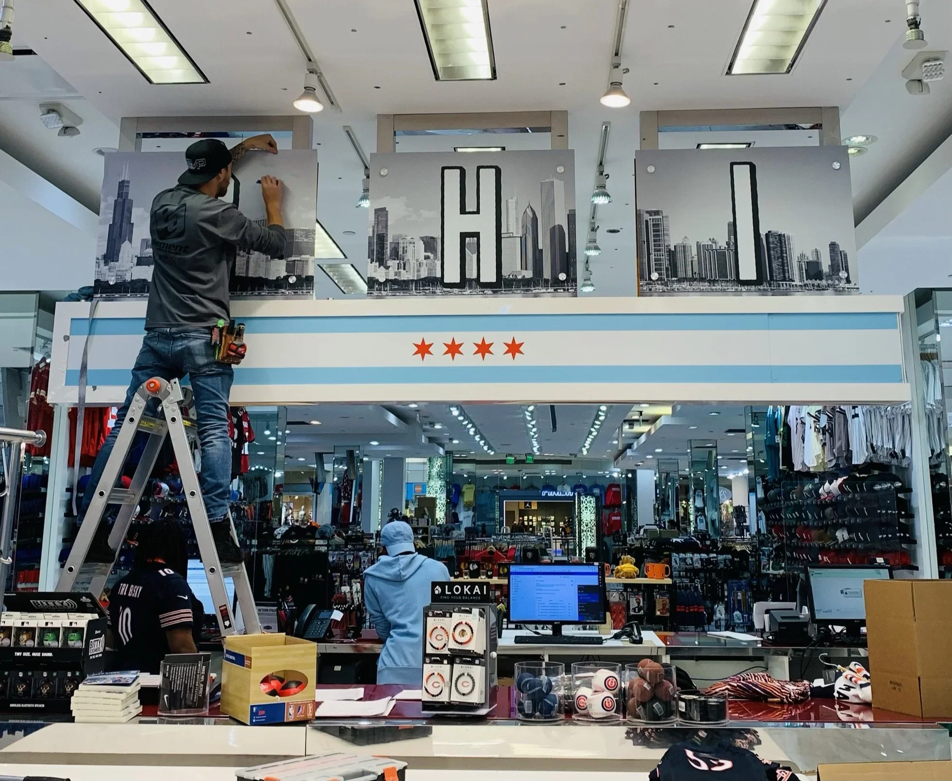 Inside a retail store with a worker on a ladder adjusting a large display with a city skyline and the word "CHICAGO" on it. The store has various items for sale including books, accessories, and clothing.
