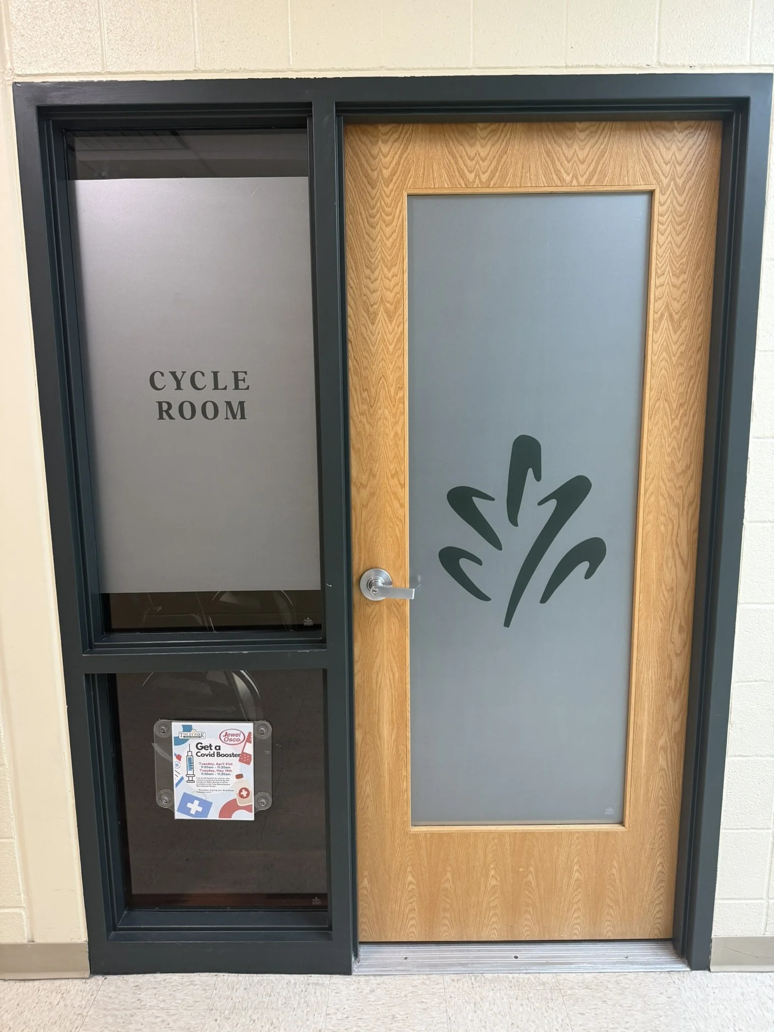 Entrance to a room with a door and window, labeled 'CYCLE ROOM' on a gray panel to the left of the door, which has a wooden frame and a graphic of a stylized plant or tree.