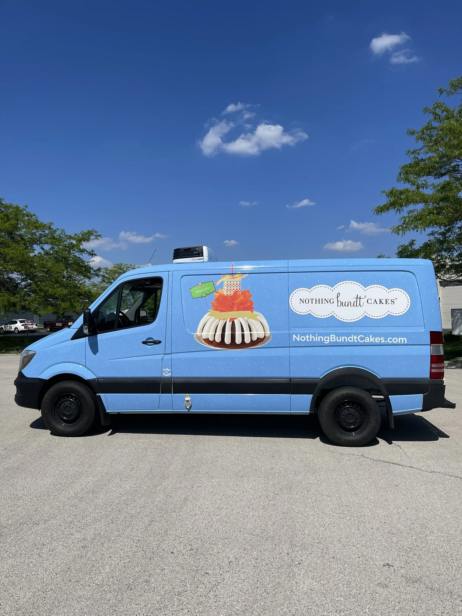 Blue delivery van with colorful cake advertisement, parked in a lot with trees and a blue sky with clouds.