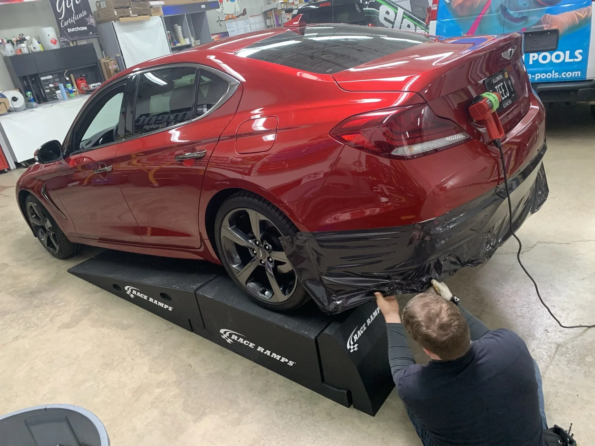 Red sports car on a Race Ramps work platform in a garage or workshop, with a man working underneath and another person inspecting from the side.