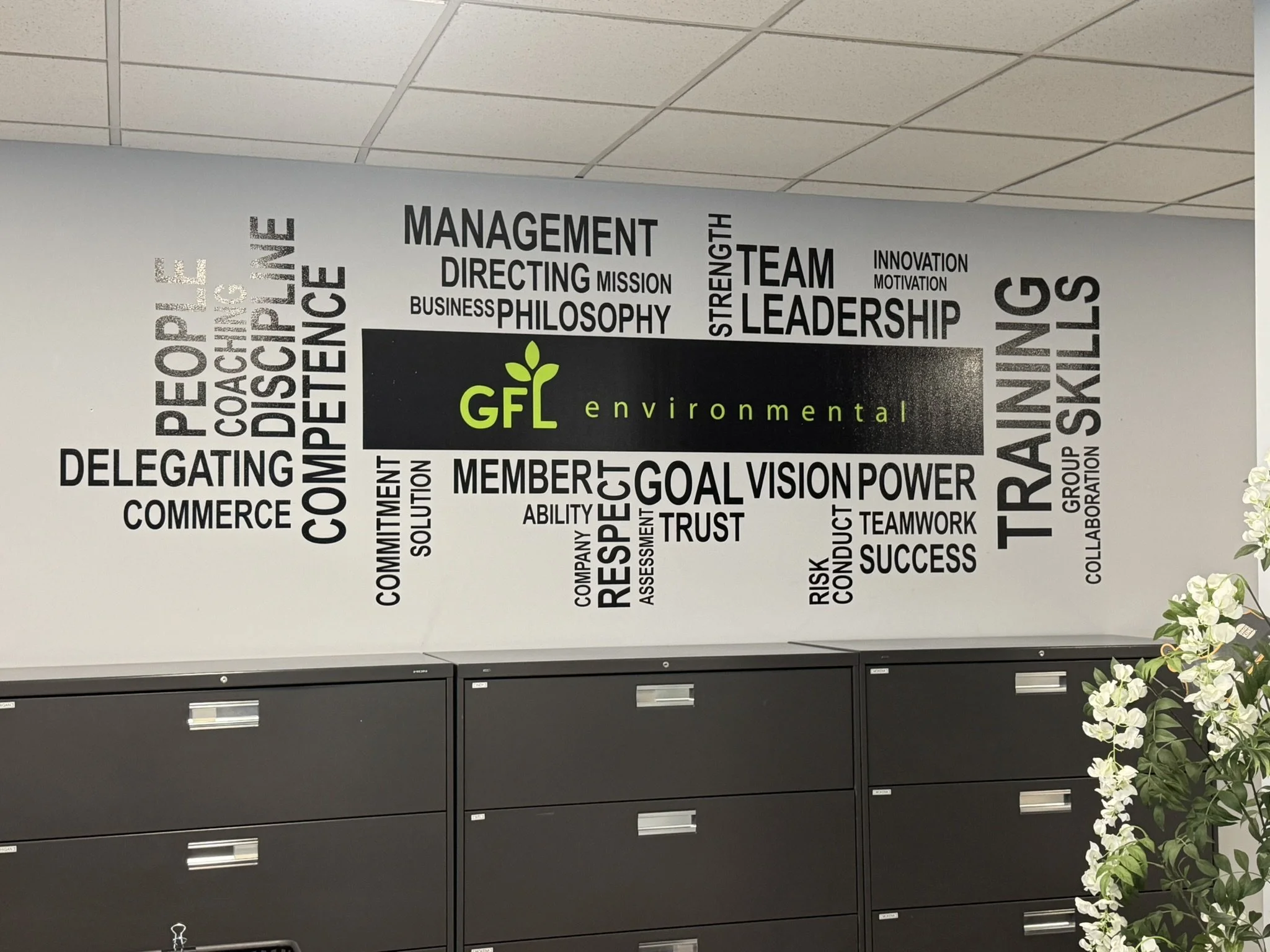 A wall decorated with words related to management, leadership, and success, with the logo and name of the company 'GFL Environmental'. There are also cabinets and a flower arrangement visible.
