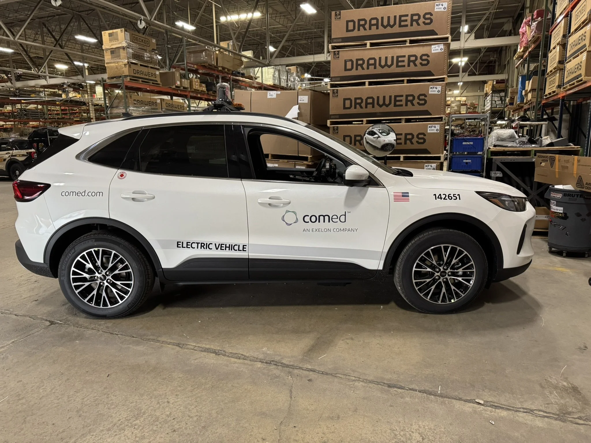 A white electric vehicle with ComEd branding parked inside a warehouse or storage facility, surrounded by stacked large cardboard boxes labeled 'DRAWSERS' and other industrial shelves.